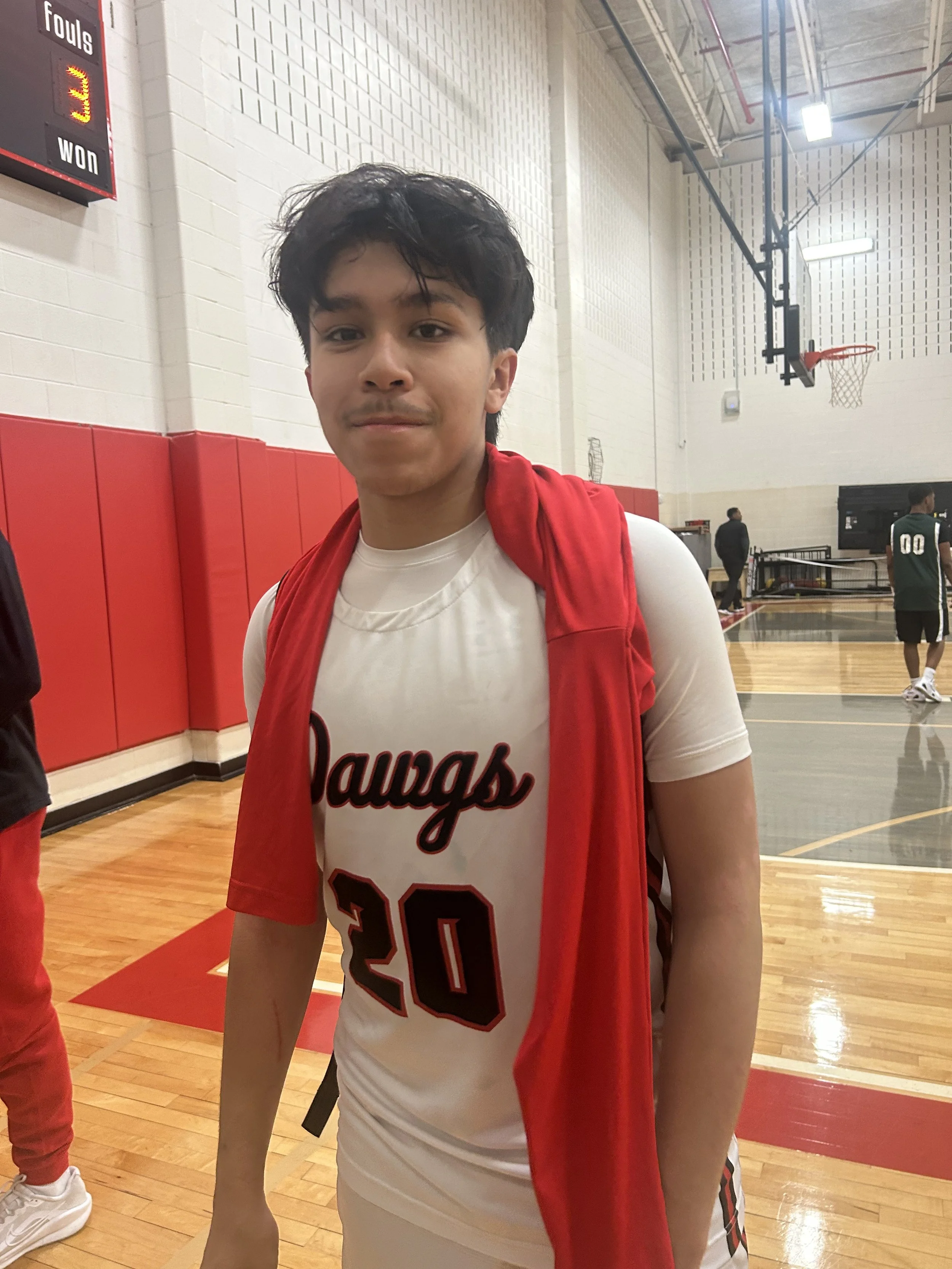 Young male basketball player in a white jersey with red and black accents, number 20, standing on a basketball court inside a gymnasium, with a red towel draped over his shoulders.
