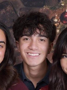 Young man with curly dark hair smiling, flanked by two women, in front of a holiday-themed background with Christmas ornaments.