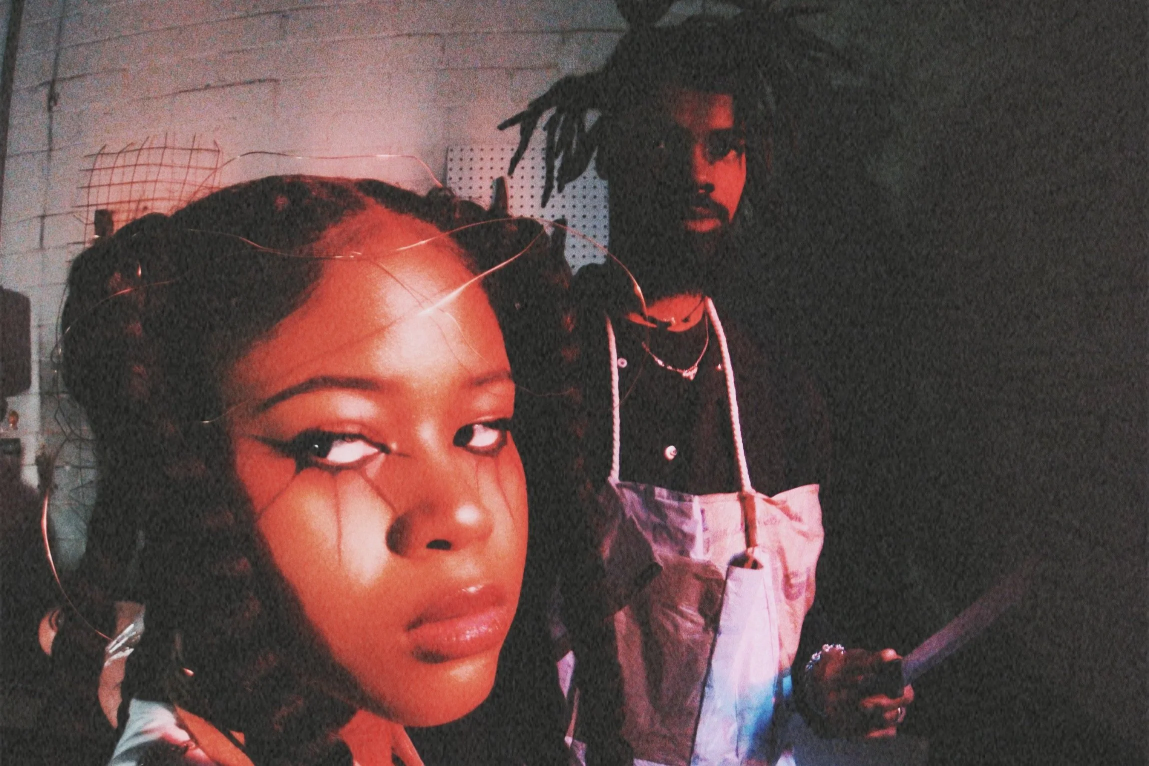 A woman with braids and makeup poses in the foreground, with a man with dreadlocks and jewelry standing behind her in a dark room with red lighting.