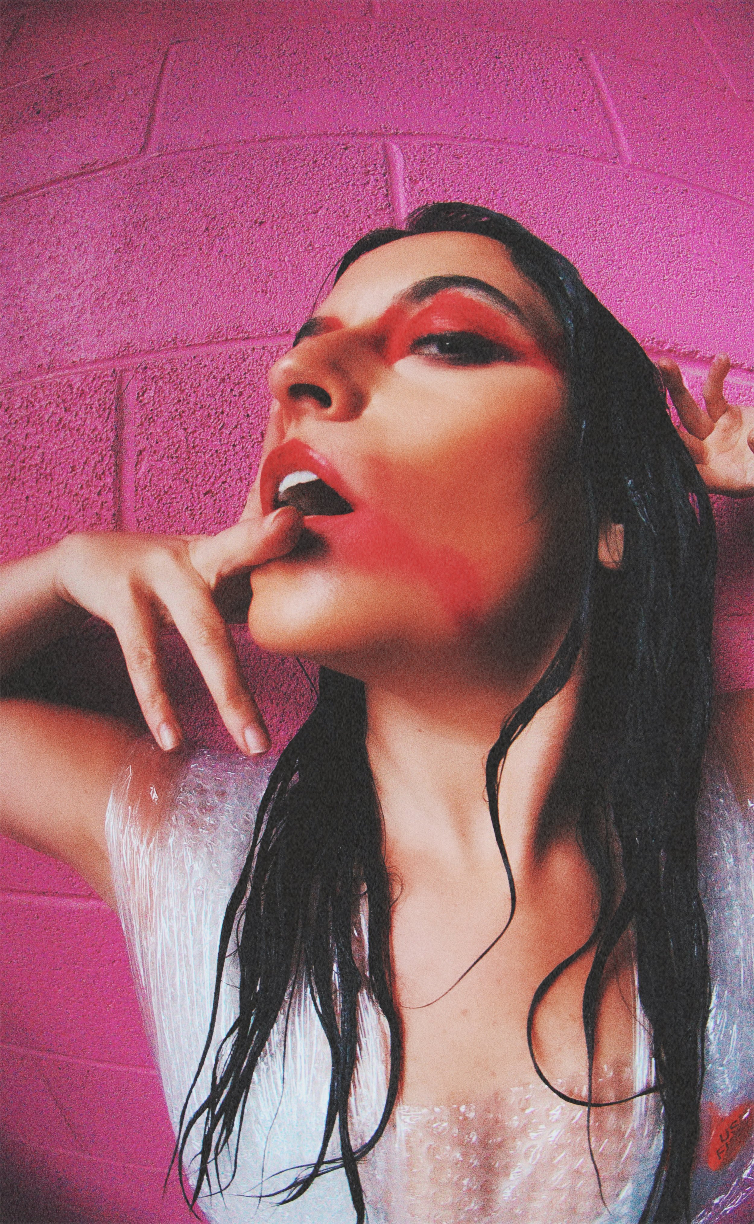 A woman with wet black hair and bold red makeup on a pink brick wall background, posing with her mouth slightly open and finger near lips.