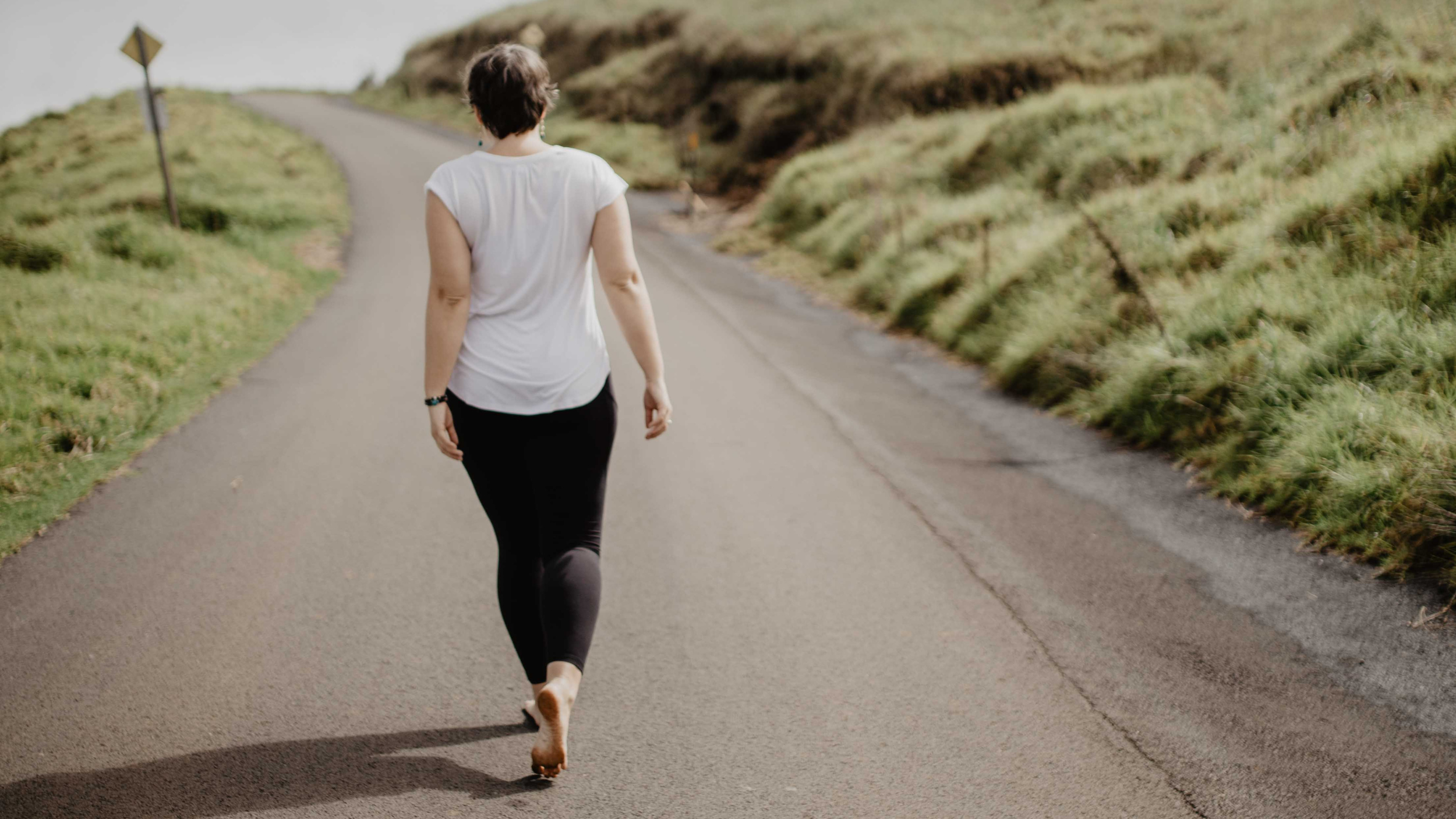 A woman walking barefoot on a rural paved road surrounded by grassy hills on a cloudy day.