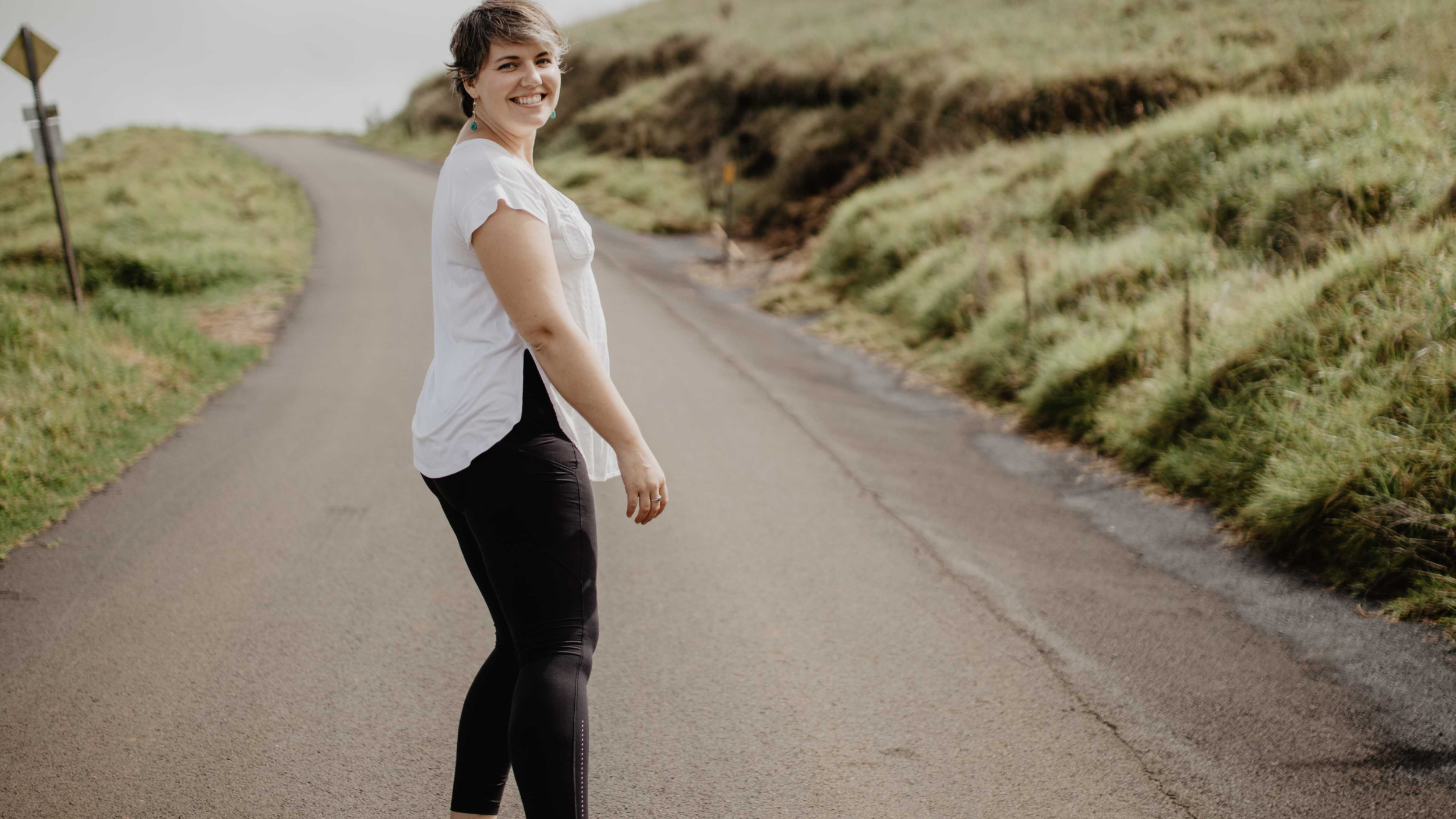 A woman with short brown hair smiling, wearing a white blouse and black pants, standing on a winding rural road with grassy hills on either side.