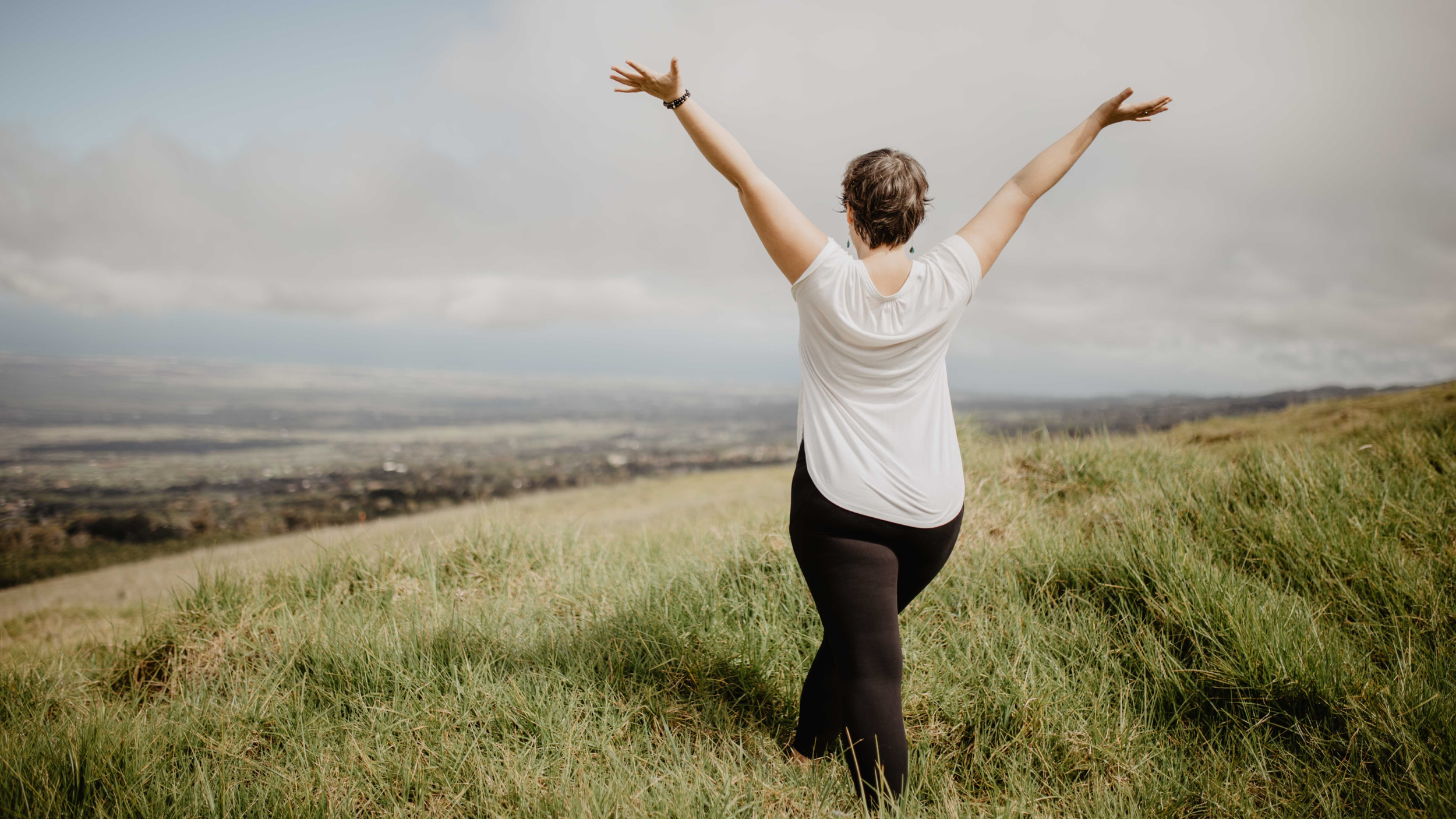 Person standing on a grassy hillside with arms raised, overlooking a cloudy landscape and distant town.