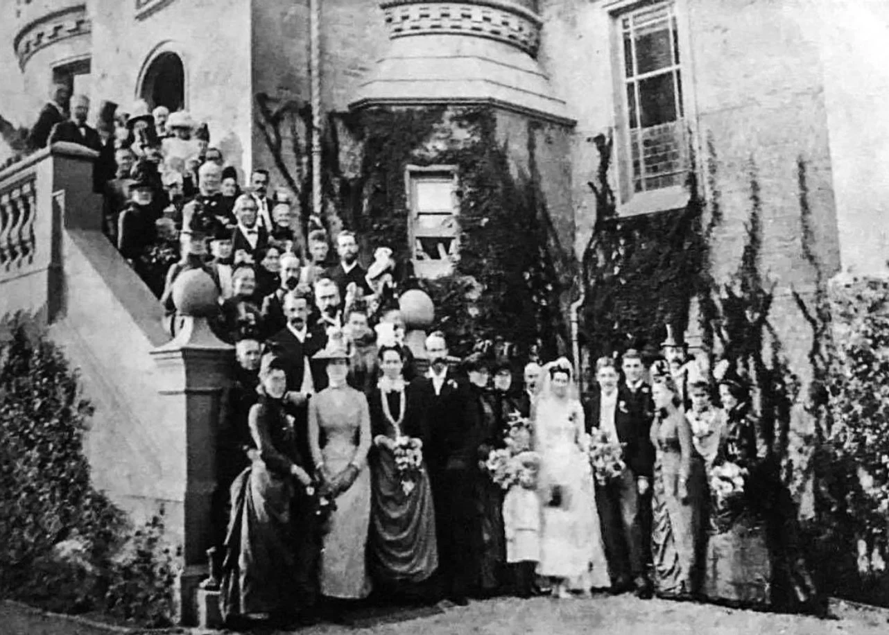 Black and white photo of a large group of people, including a bride and groom, gathered on steps and in front of a large house, celebrating a wedding.