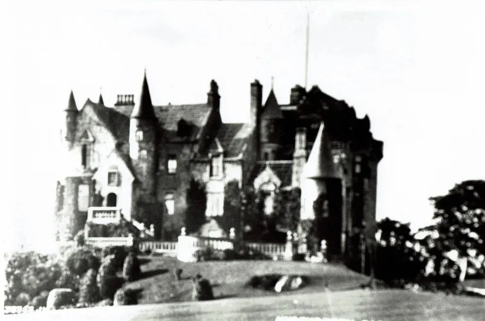 Black and white photograph of a large, old castle with tall towers and turrets, set on a hill with trees and shrubs in the foreground.