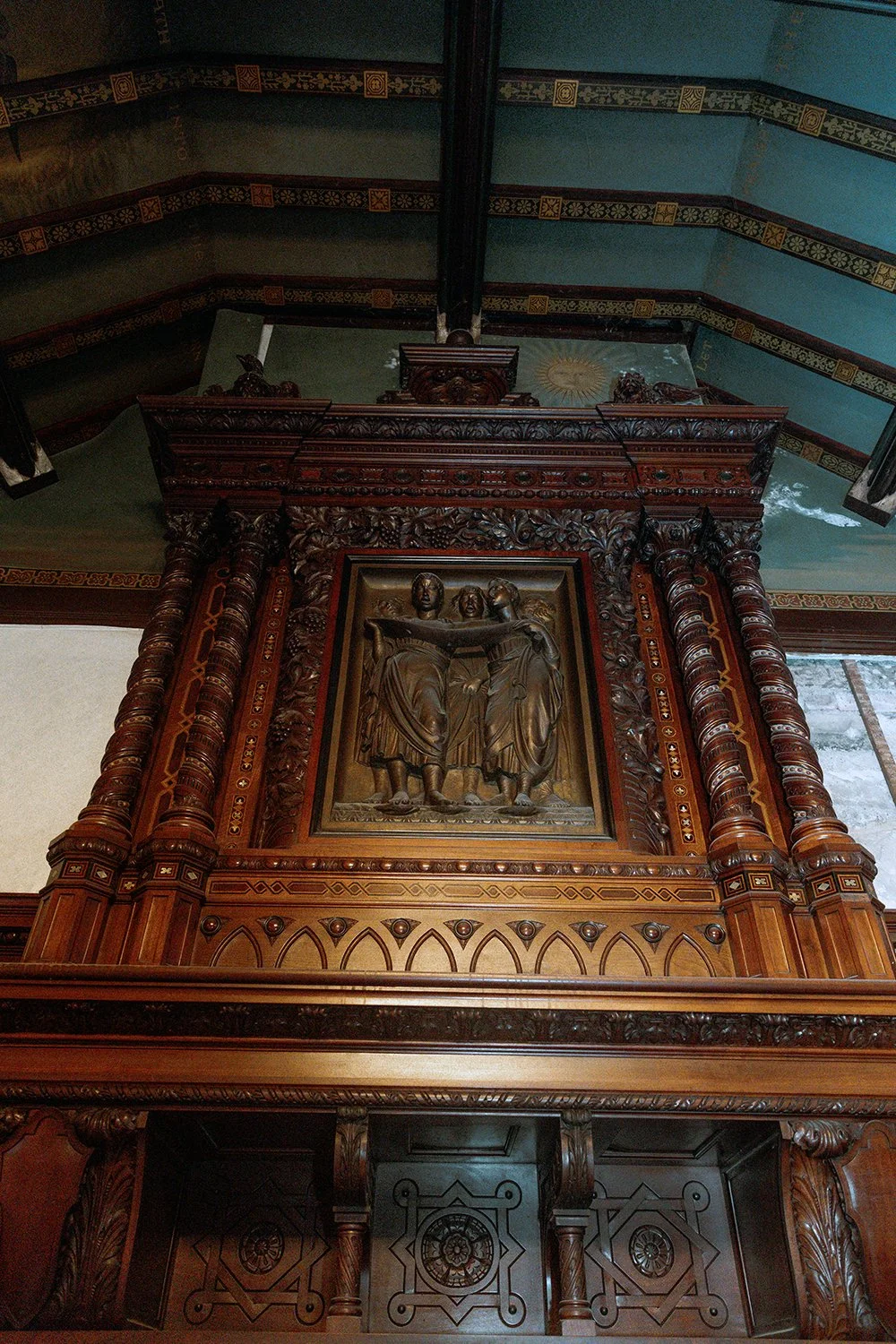 A carved wooden panel depicting two figures in a religious scene, surrounded by ornate woodwork and decorative columns, inside a church or cathedral.