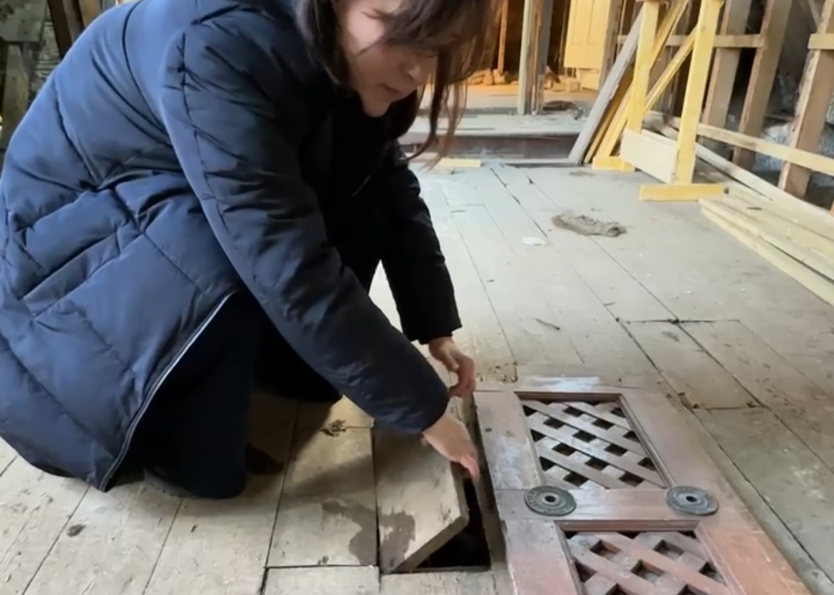 A woman in a dark jacket crouches down on a wooden floor, lifting a wooden board near a floor vent in a construction site.
