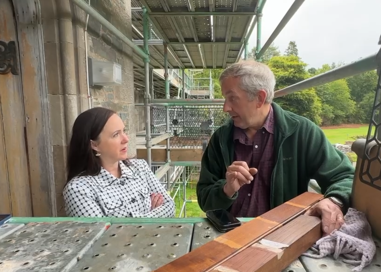 A woman with dark hair and a checkered shirt stands with arms crossed, listening to an older man with gray hair and a green jacket who is speaking and gesturing with his hand, outside near a construction scaffold.