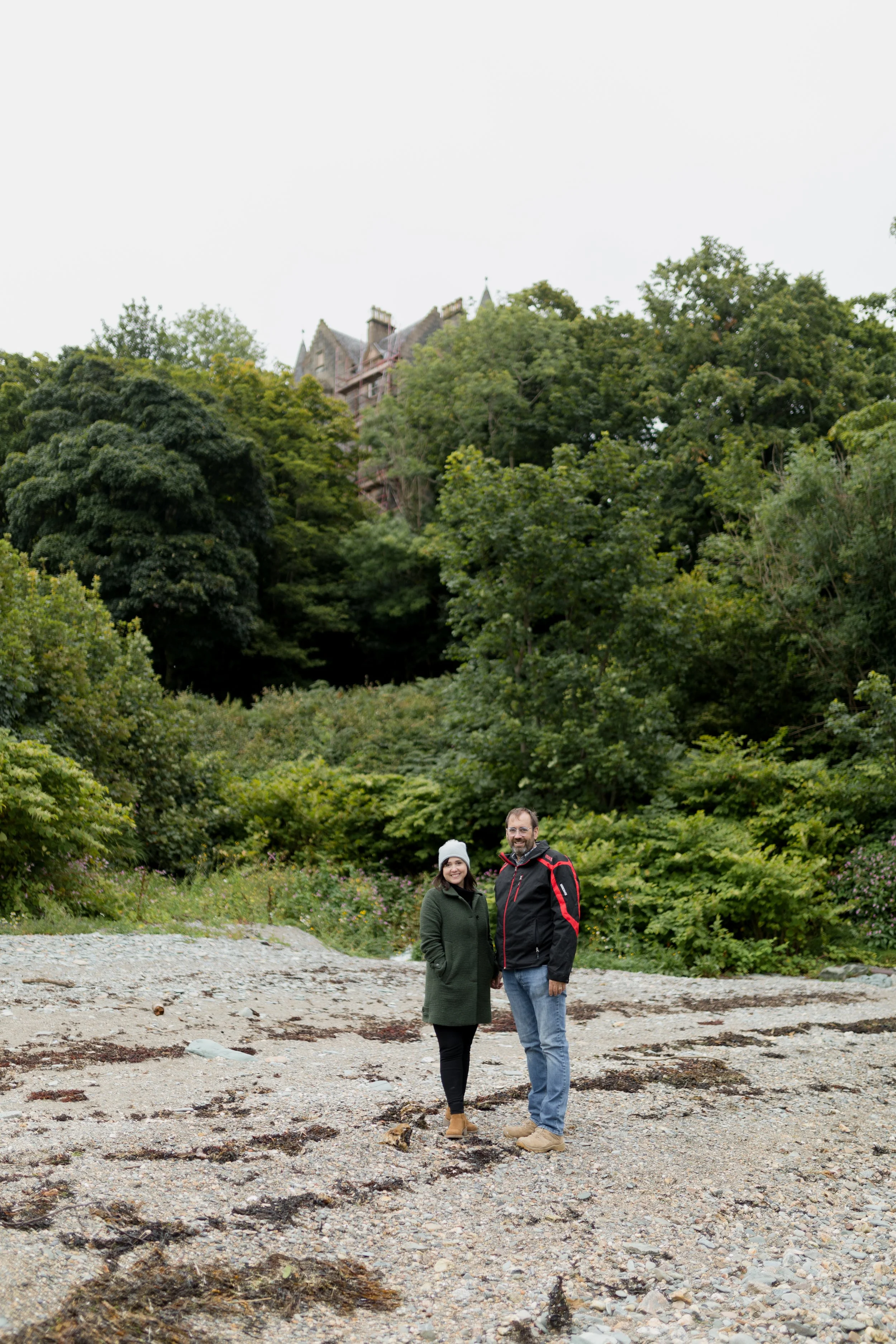 A couple standing on a rocky shore with dense green trees in the background and a castle-like building partially visible on a hilltop. The woman is wearing a green coat and a grey beanie, and the man is wearing a black jacket with red accents and jeans.