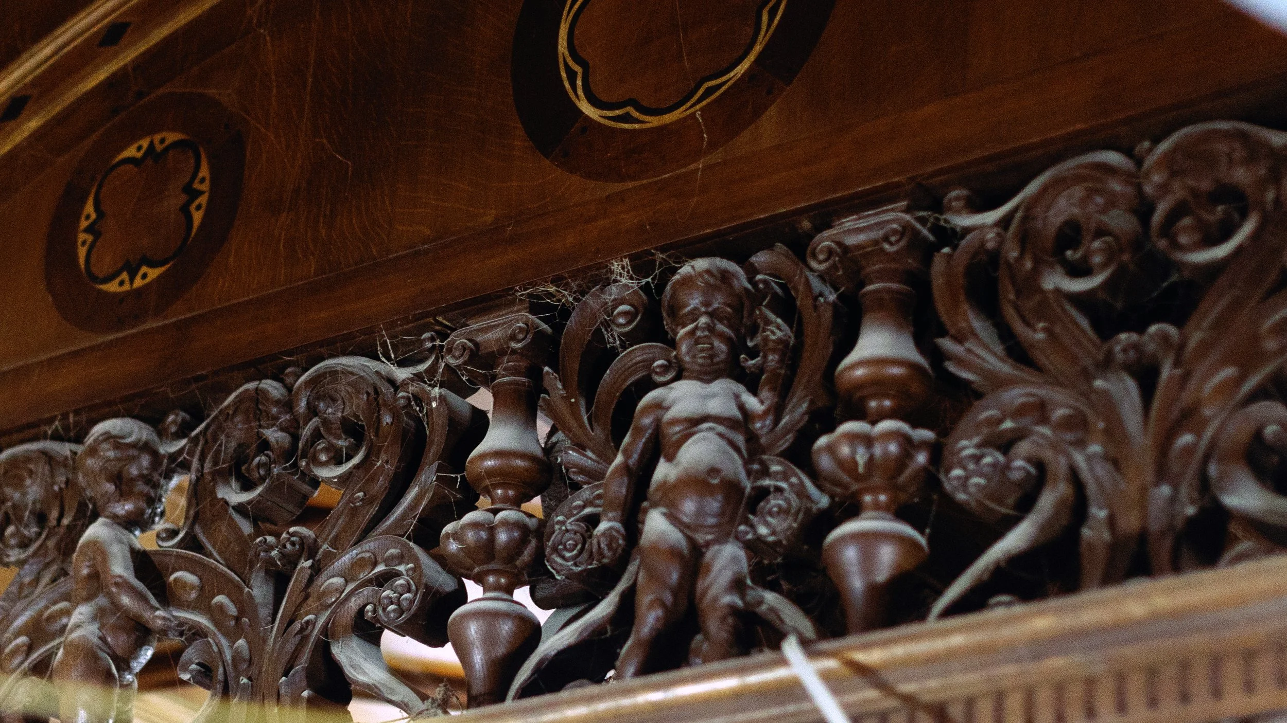 Detailed close-up of ornate carved wood furniture with cherubic children and decorative scrolls, some cobwebs present.