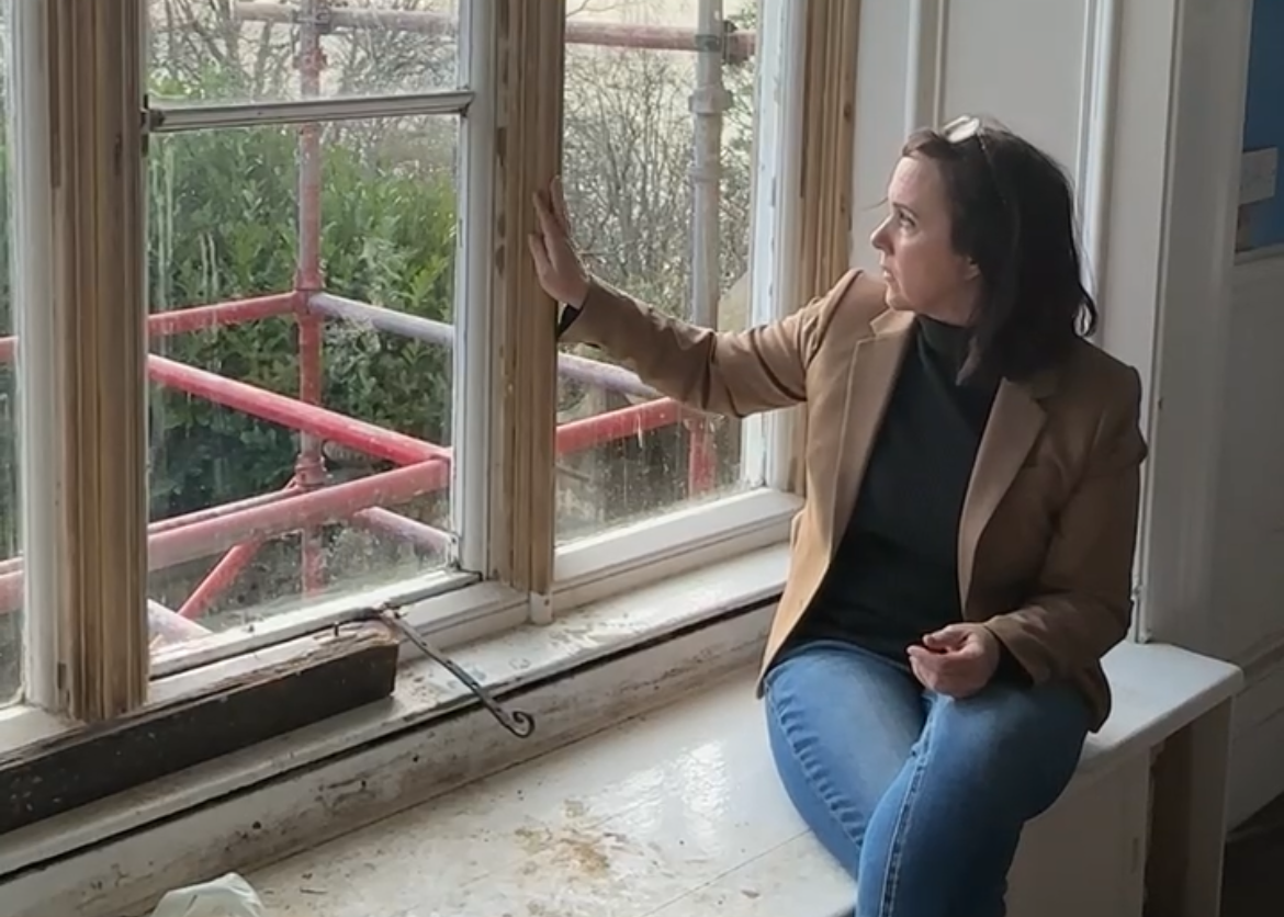 A woman sitting on a windowsill with her hand on the window frame, looking outside.