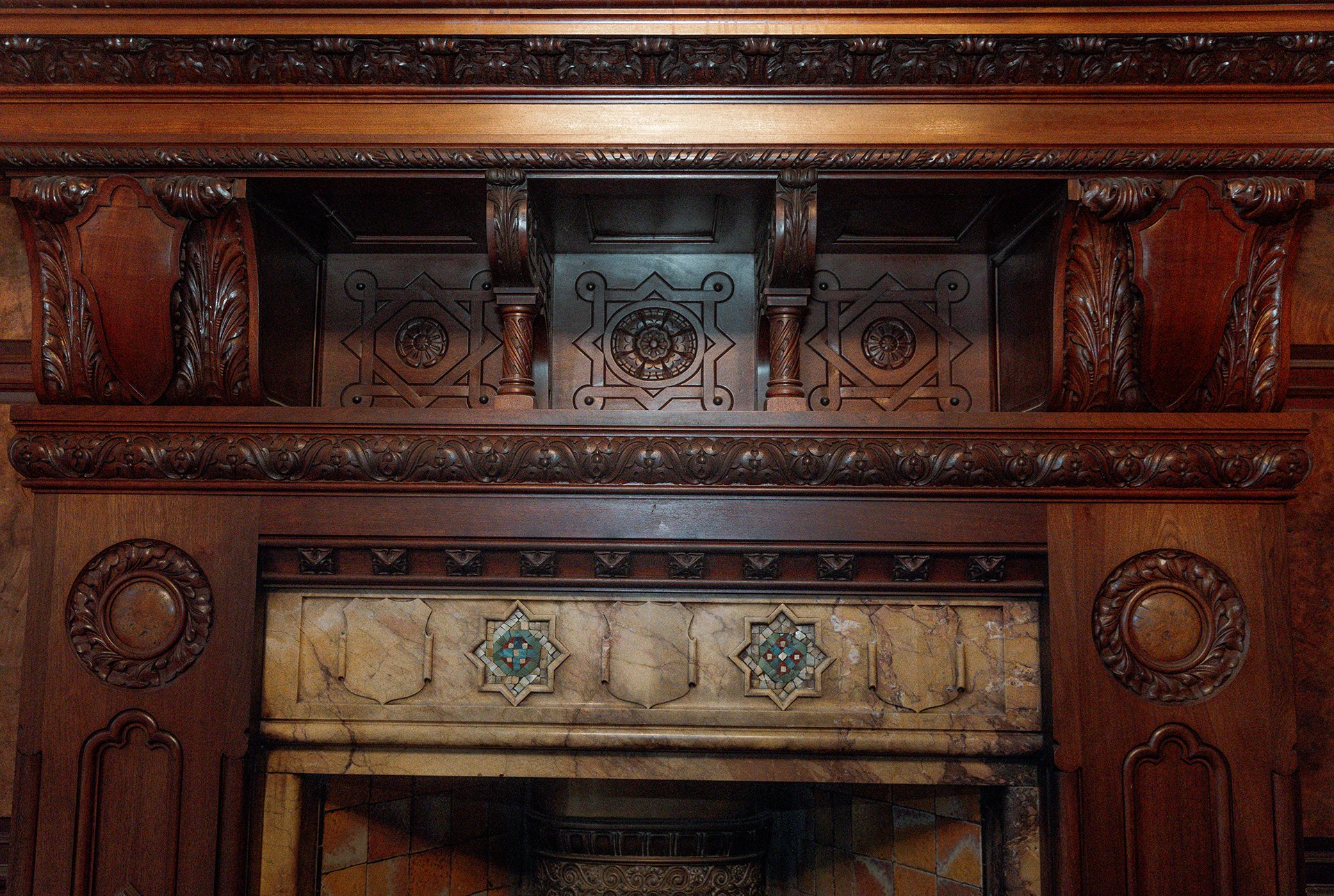 Close-up view of an ornate, dark wooden fireplace mantel with intricate carvings, columns, and a decorative tile inlay with mosaic patterns.