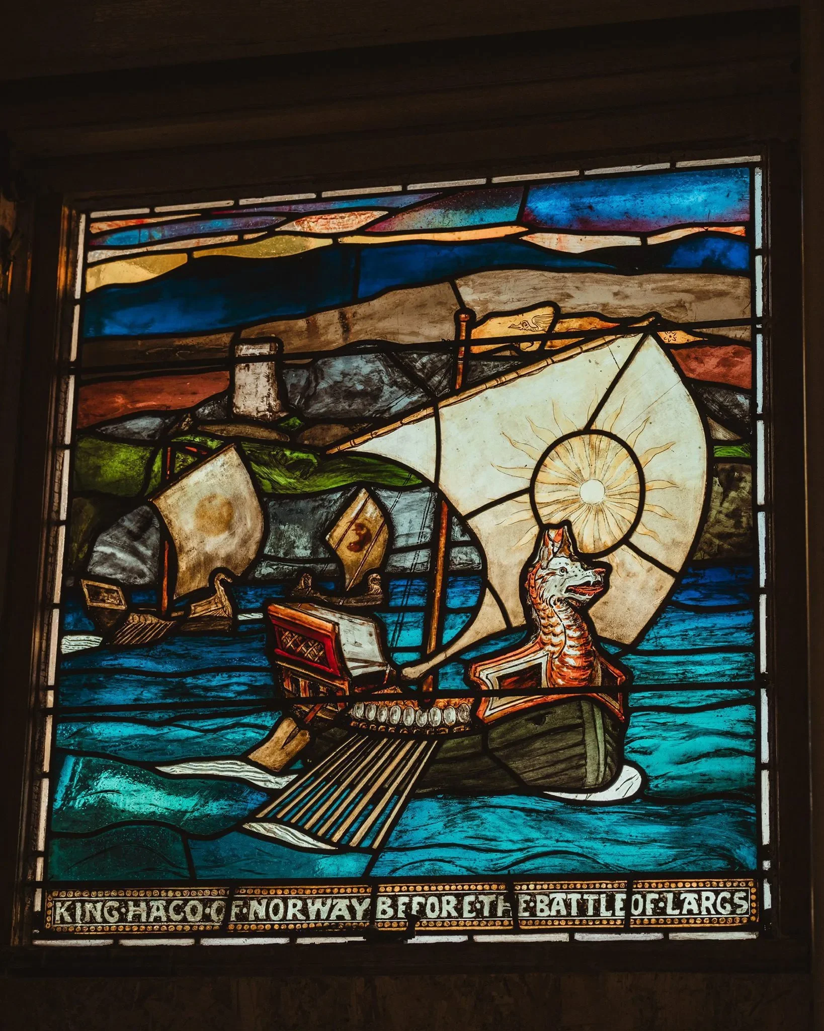Colorful stained glass window depicting a Viking ship with a dragon figurehead sailing on the water, under a sunrise or sunset, with the text 'King Haco of Norway Before the Battle of Largs' at the bottom.