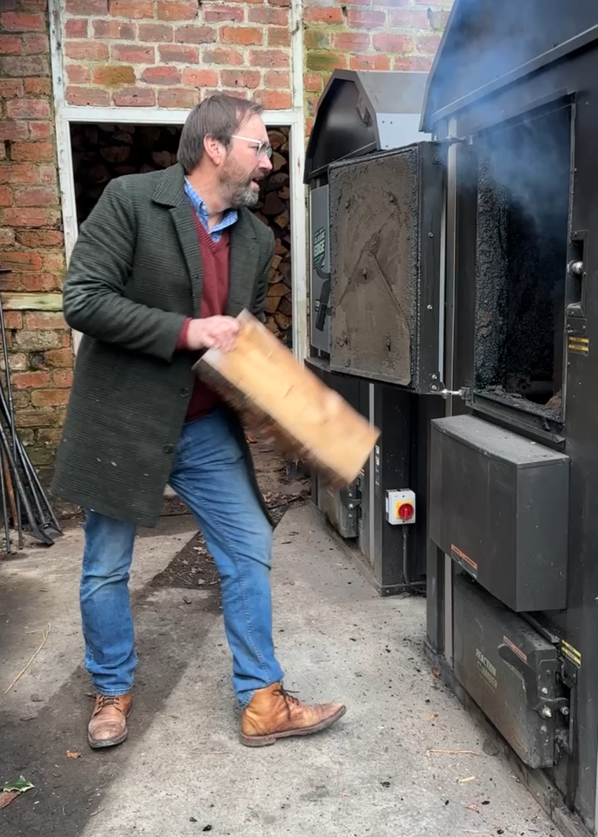 A man with glasses, a beard, and wearing a dark coat is throwing a piece of wood into a fire in an outdoor wood kiln, with brick wall and woodpile in the background.