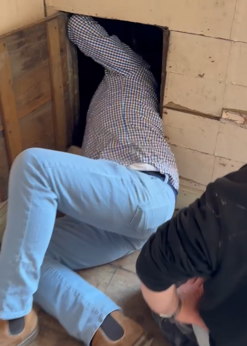 Someone crouched down inspecting a crawl space in a wooden wall. Another person is kneeling nearby.