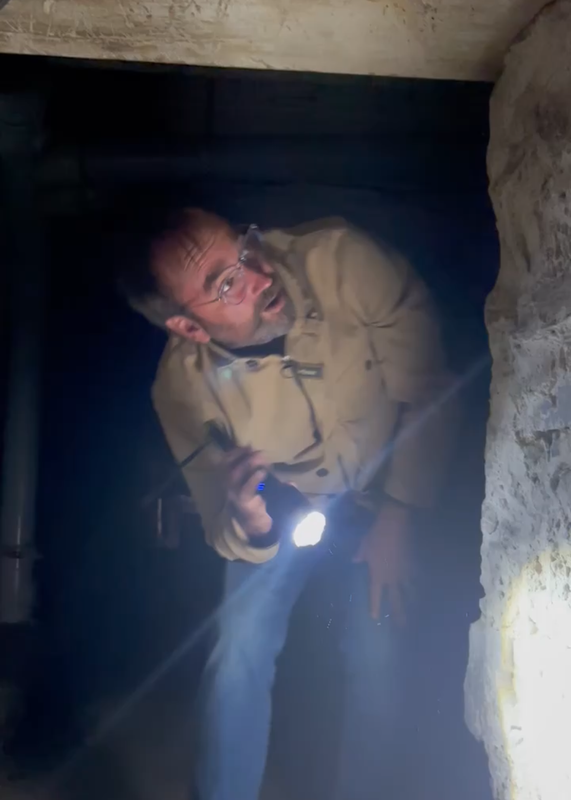 A man in a tan jacket, wearing glasses, holding a flashlight in a dark space and looking back over his shoulder.
