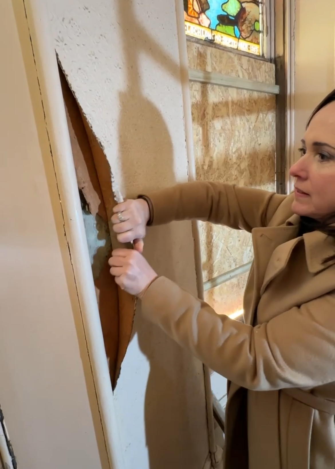 Woman in a tan coat pulling down damaged drywall from a wall, revealing insulation and framing behind.