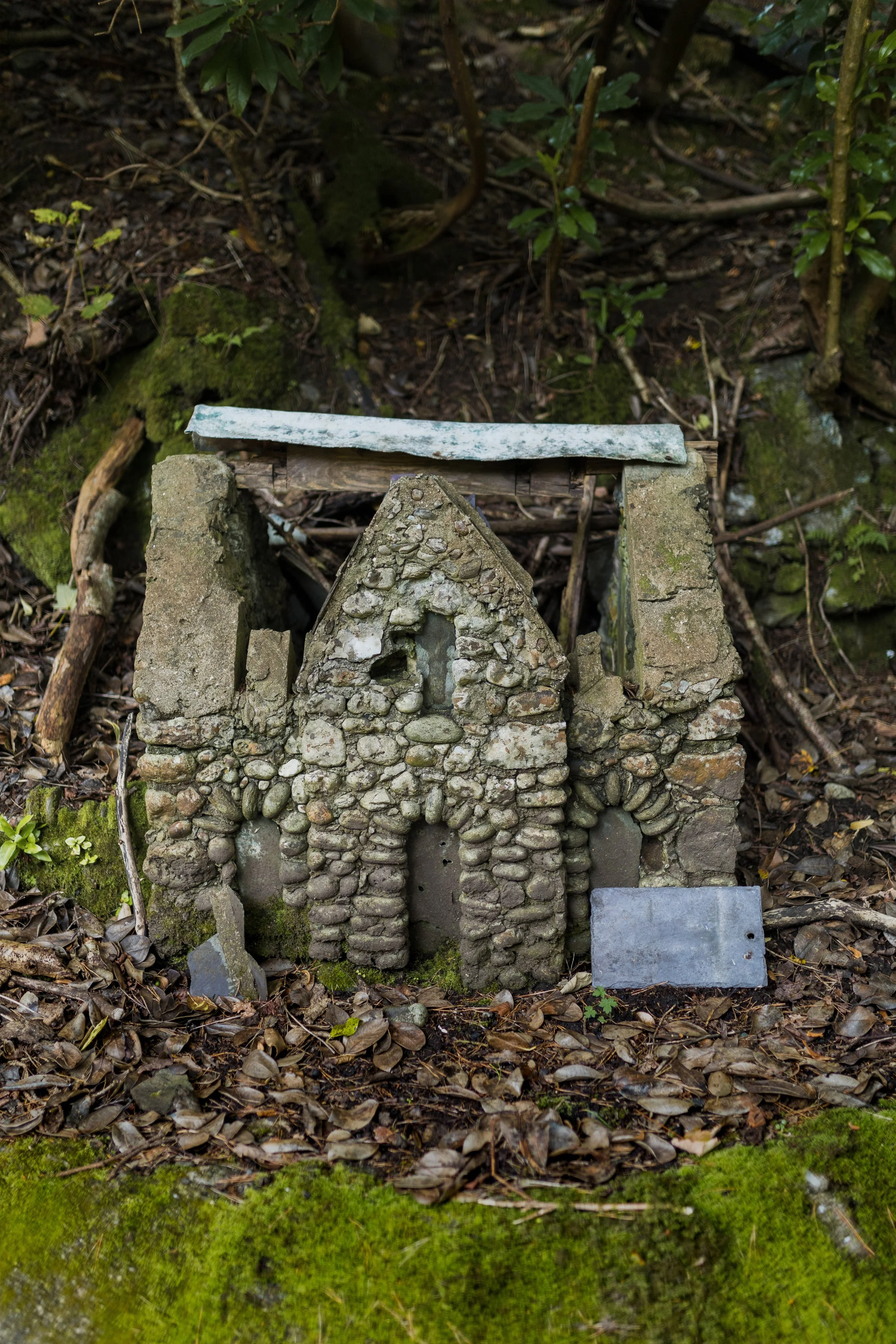 Small stone and concrete model of a church with a metal roof, situated on forest floor surrounded by leaves and moss.