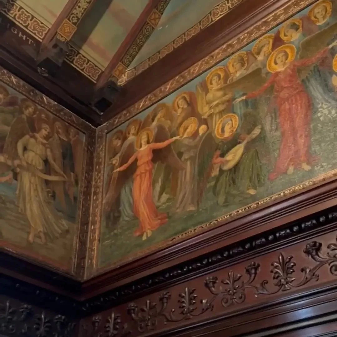 A corner of a room with elaborate wooden paneling and a detailed religious mural painting depicting angels and a figure with a halo, possibly in a historic church or cathedral.