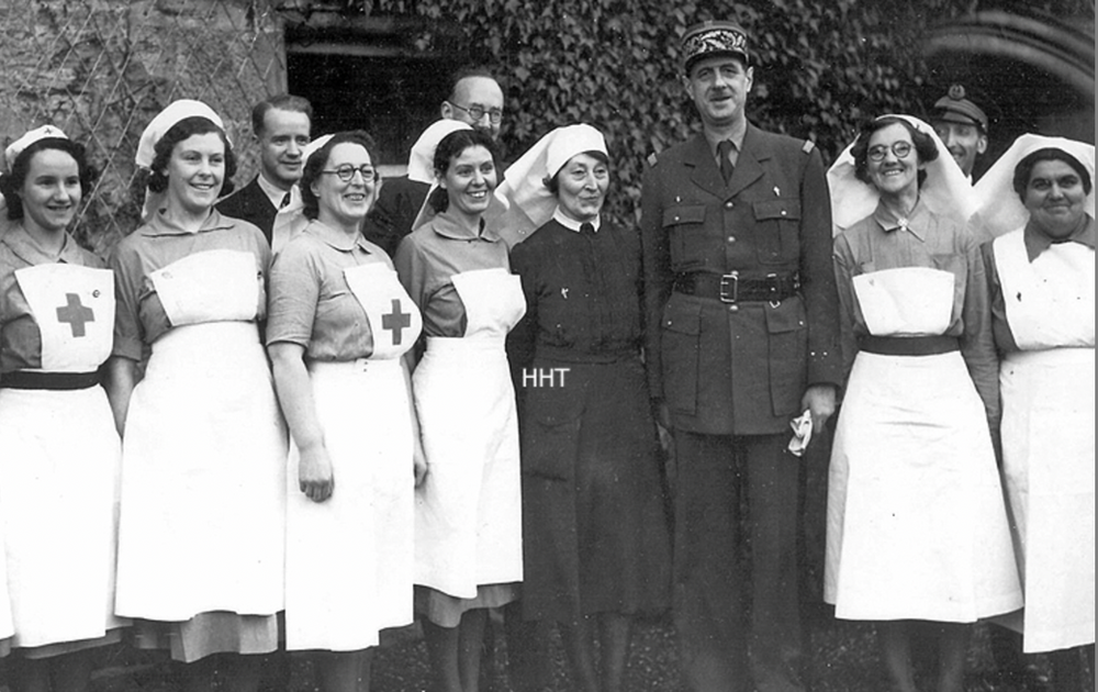 Historical black and white photo of nurses in uniforms with nurses' caps, alongside a man in a military uniform, standing outdoors in front of a wall covered in vines.