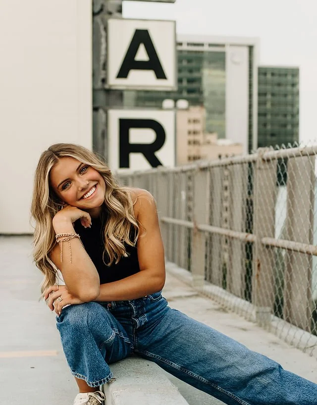 A young woman with wavy blonde hair, sitting on a parking garage ledge, smiling, wearing a black sleeveless top, blue jeans, and sneakers, with a cityscape background and large blocks with the letters A and R in the background.