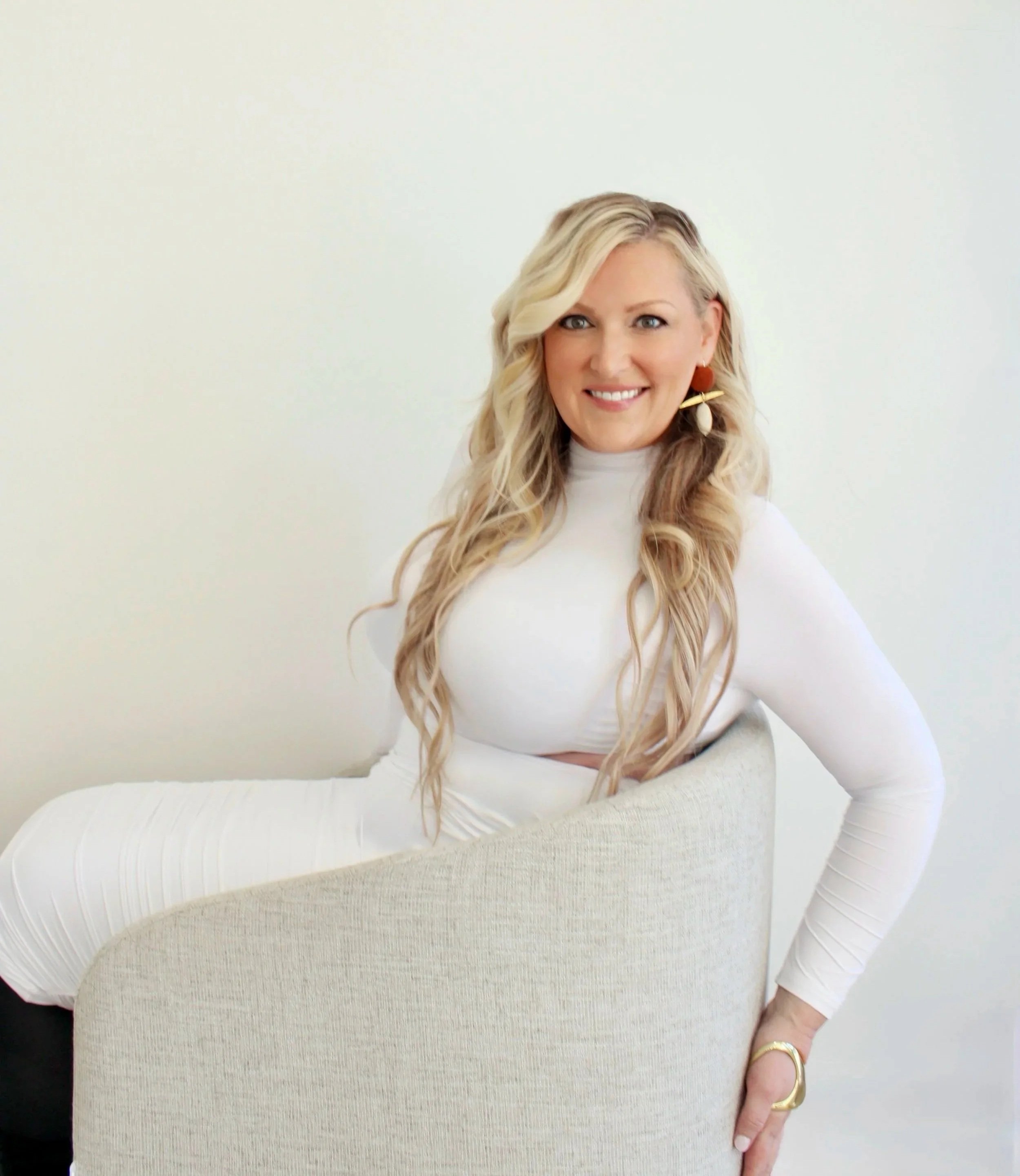 A woman with long blonde hair, wearing a white long-sleeve dress, sitting on a beige chair against a plain white background, smiling.