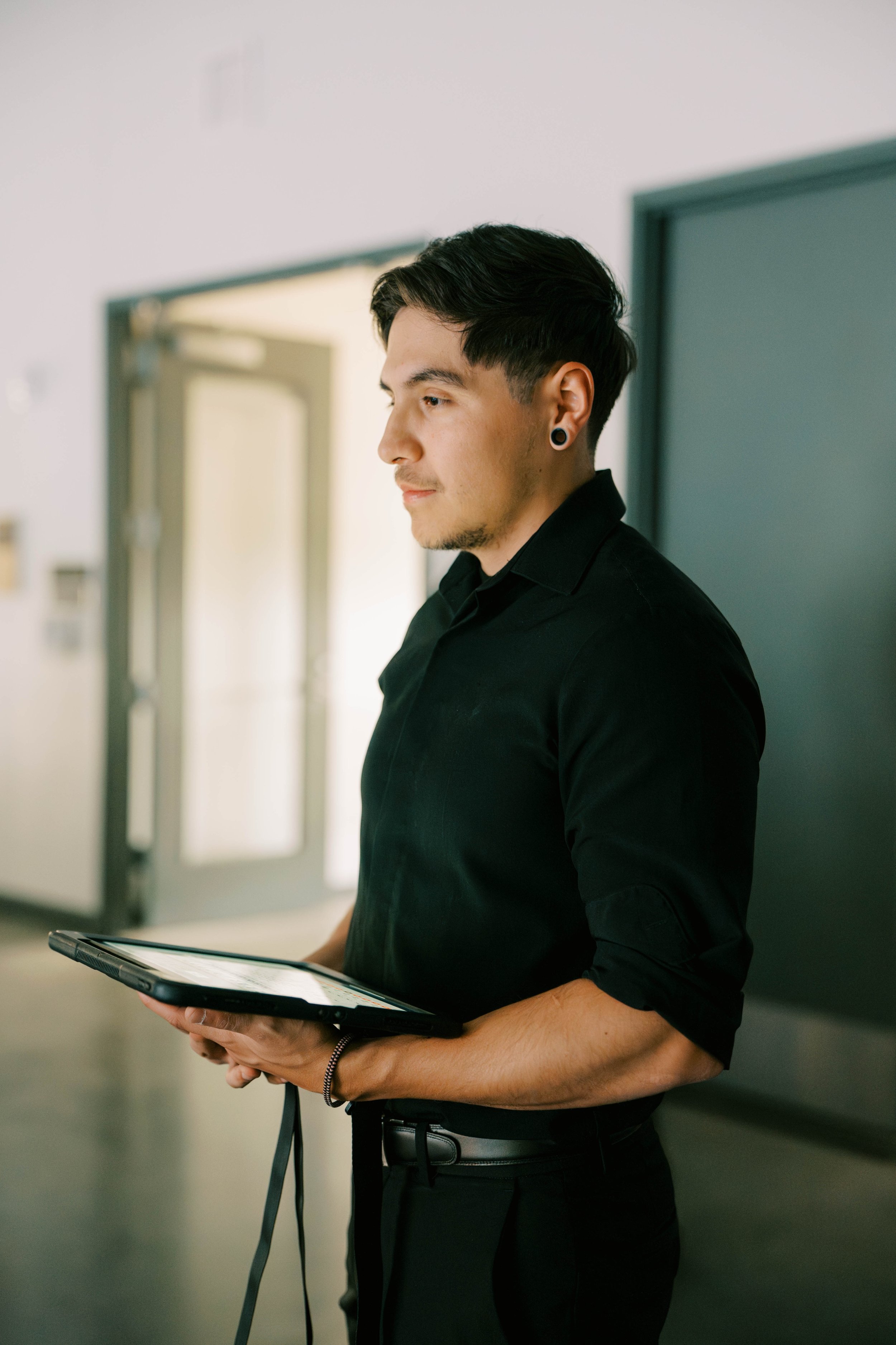 A young man with dark hair, earrings, and dressed in black, standing indoors and looking at a tablet.