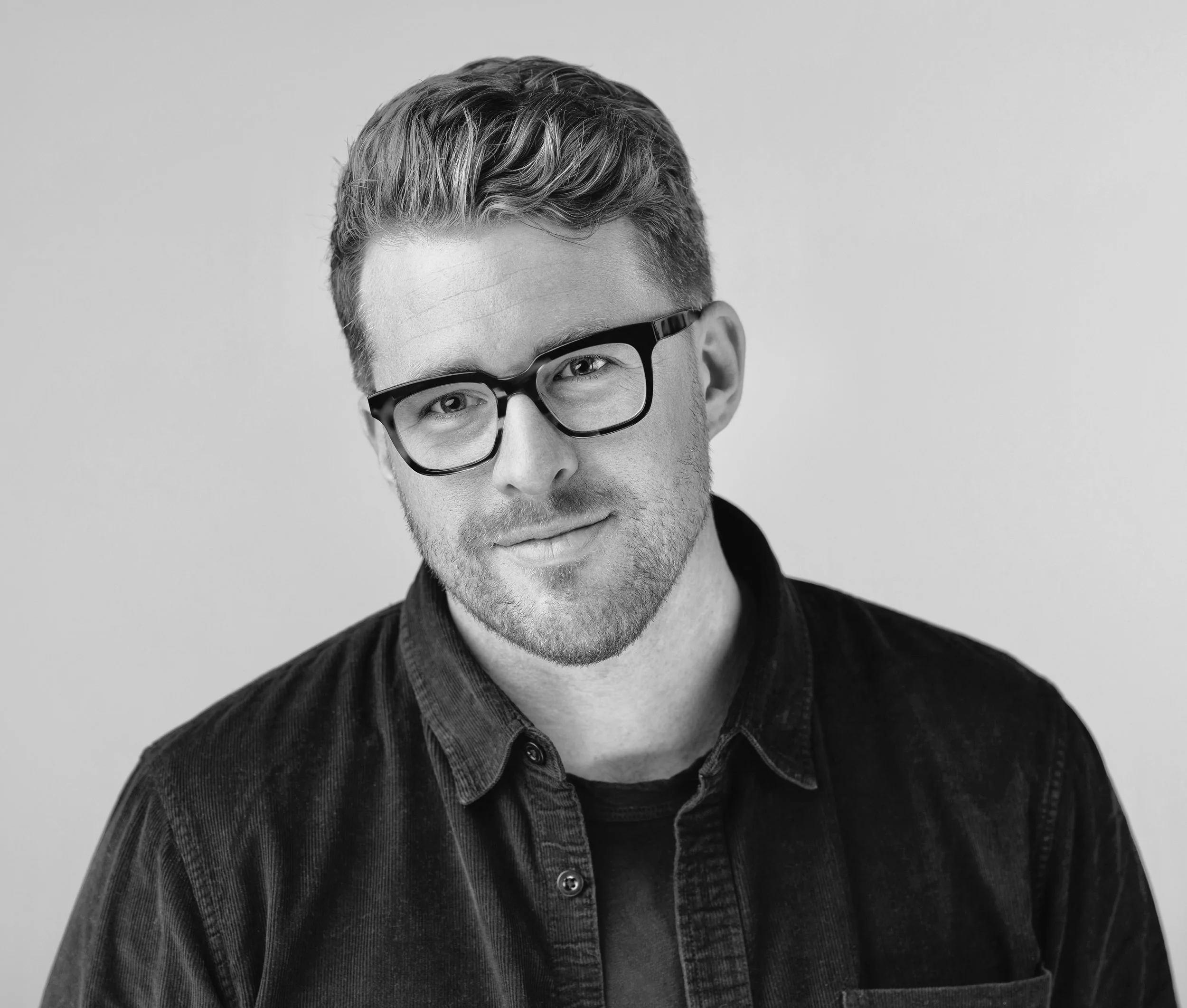 Black and white portrait of a young man with glasses, short hair, and a slight smile, wearing a dark shirt against a plain background.