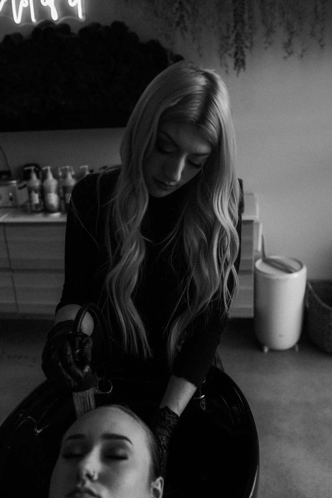 Hair stylist with long blonde hair washing a client's hair at the shampoo bowl.