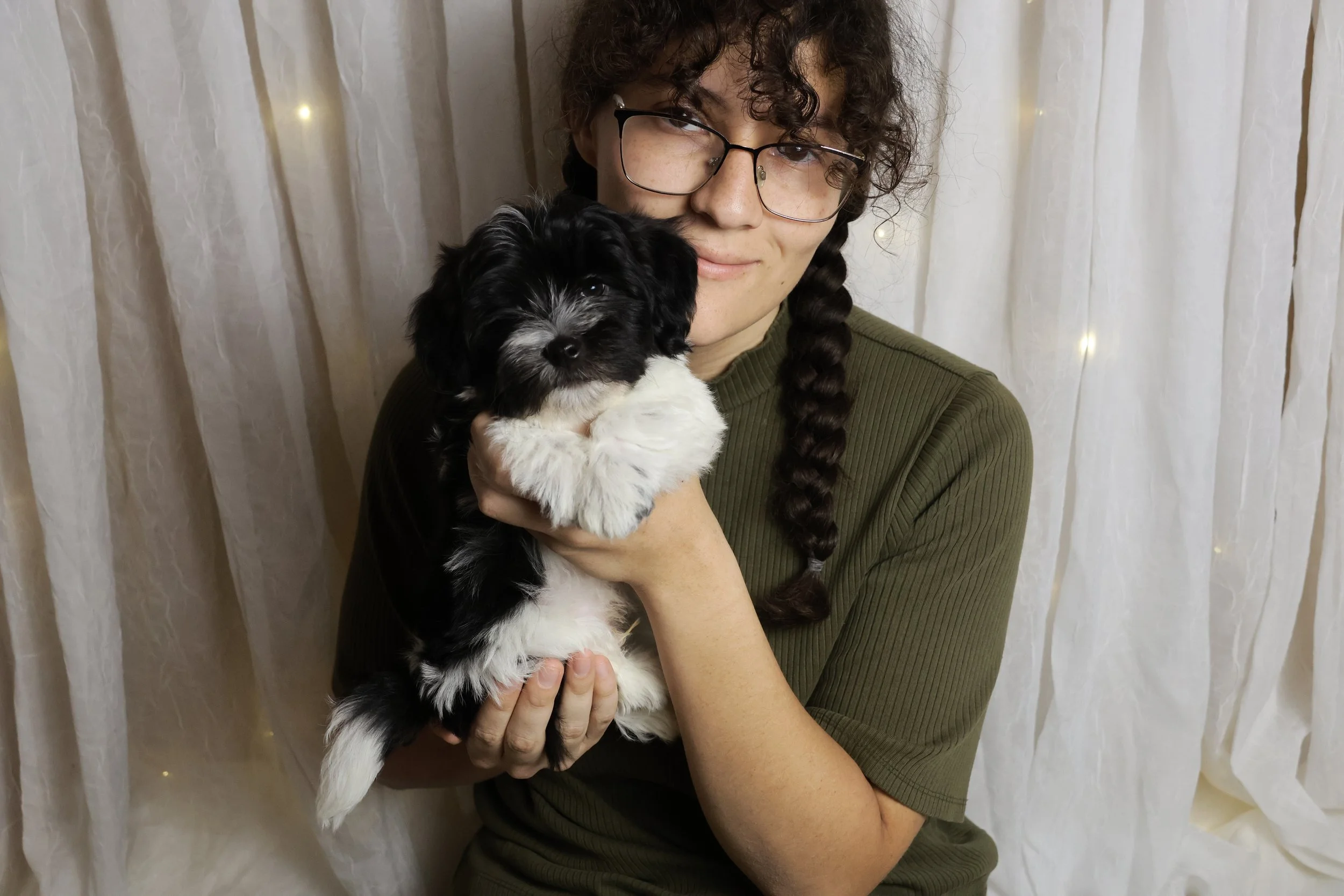 A woman with glasses and a braid holding a black and white puppy against a cream-colored curtain backdrop.