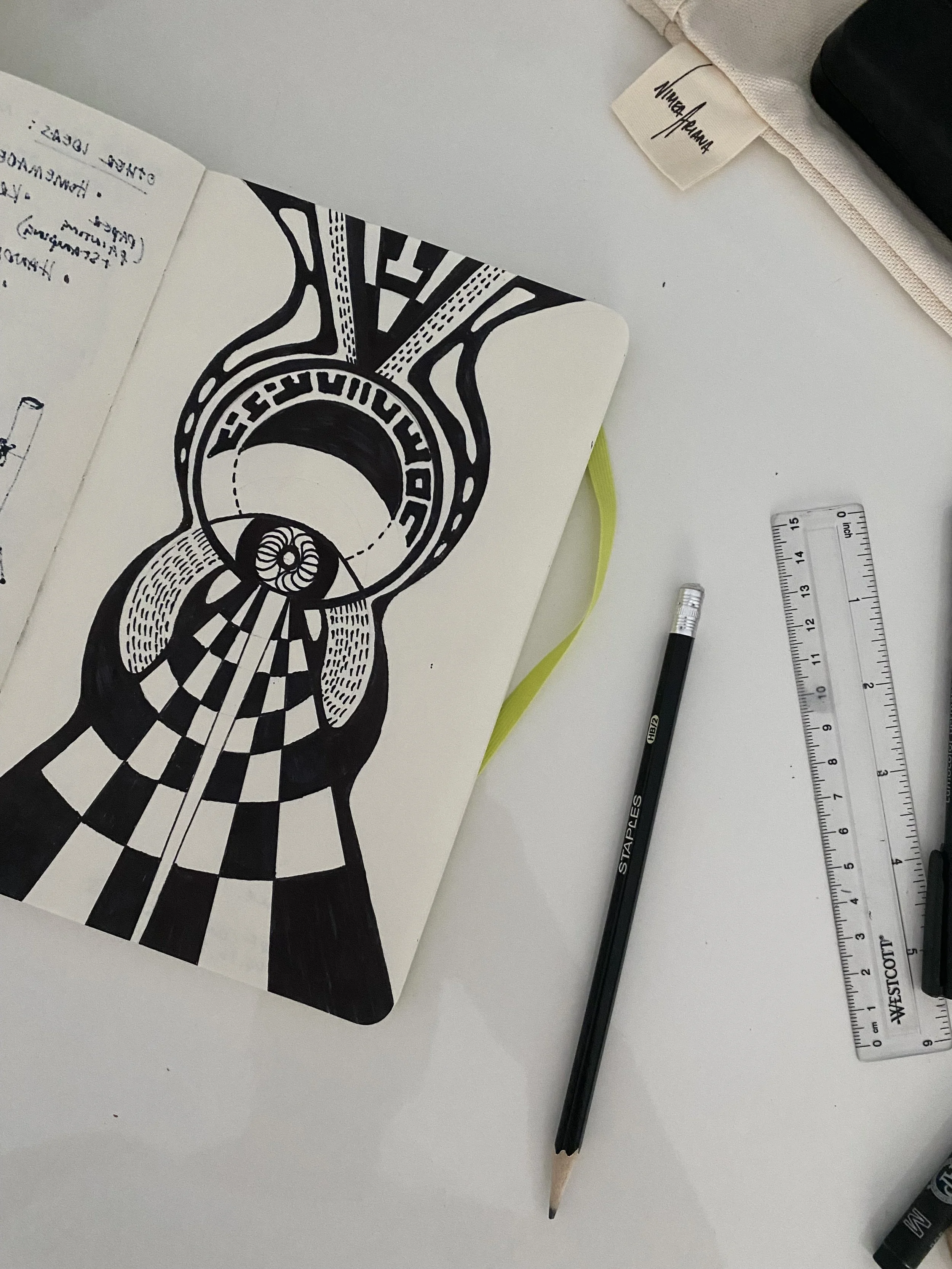 Open sketchbook with black and white abstract geometric drawings, a black pencil, a clear ruler, and other stationery items on a white desk.