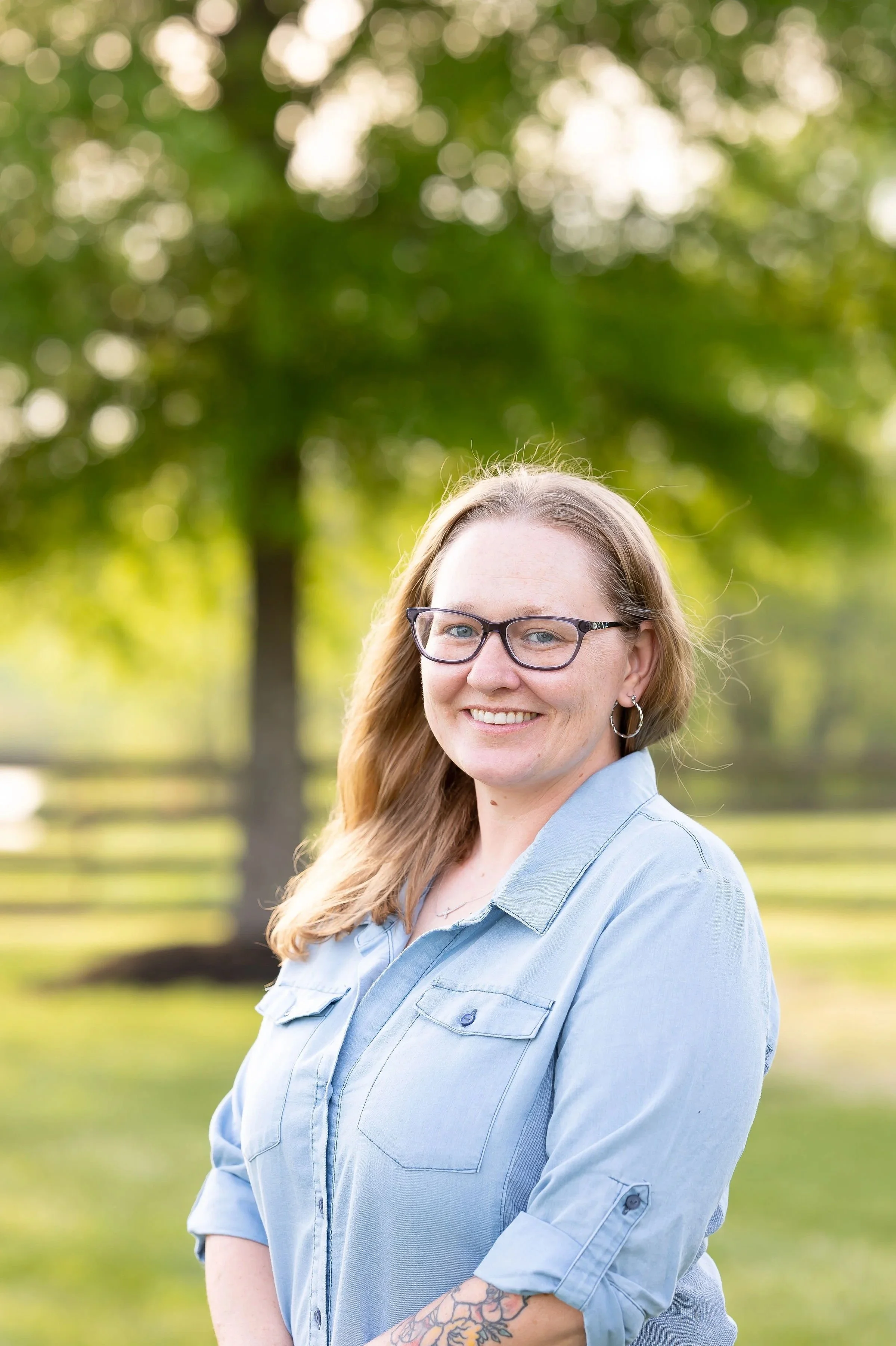 A woman wearing glasses and a light blue shirt, smiling outdoors with trees and a fence in the background.