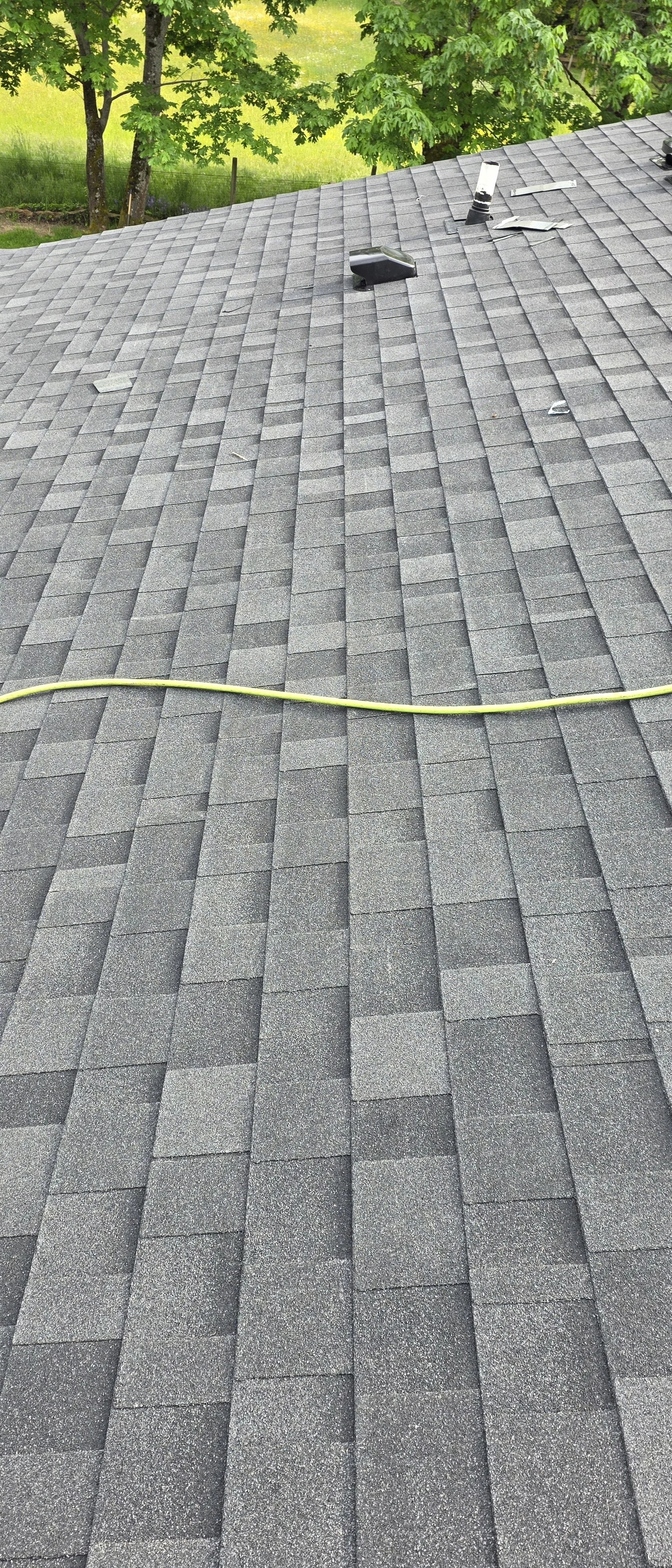 View of a gray shingled roof with tools and a yellow cord on top, with green trees in the background.
