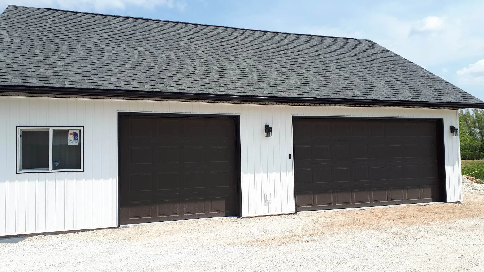 A modern two-car garage with white siding, black garage doors, and a gray shingle roof under a partly cloudy sky.