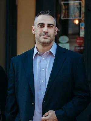A man with short dark hair and light skin wearing a dark blazer and light blue shirt standing outdoors in front of a building.