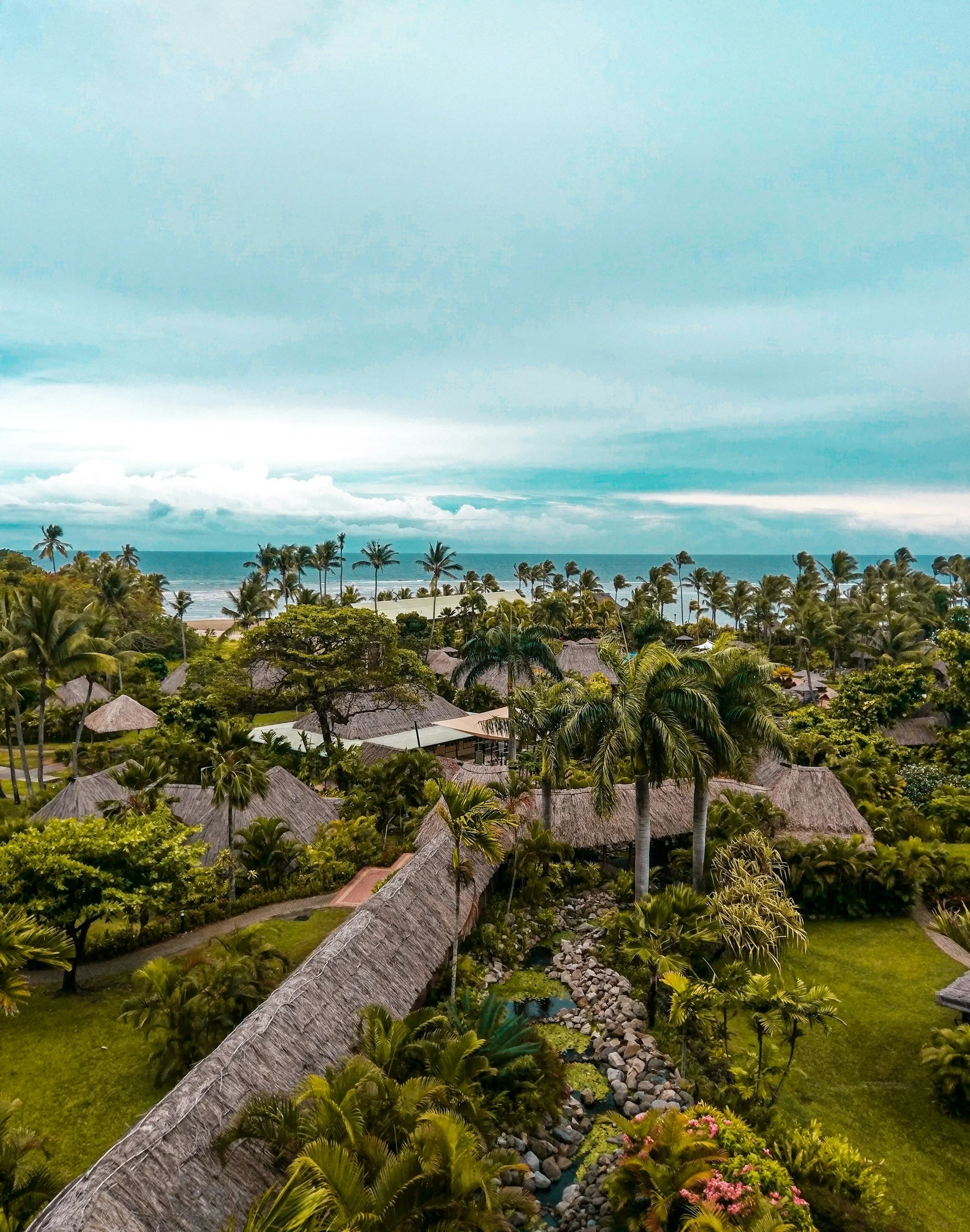 Tropical resort landscape with palm trees, thatched-roof buildings, and ocean in the background under a cloudy sky.