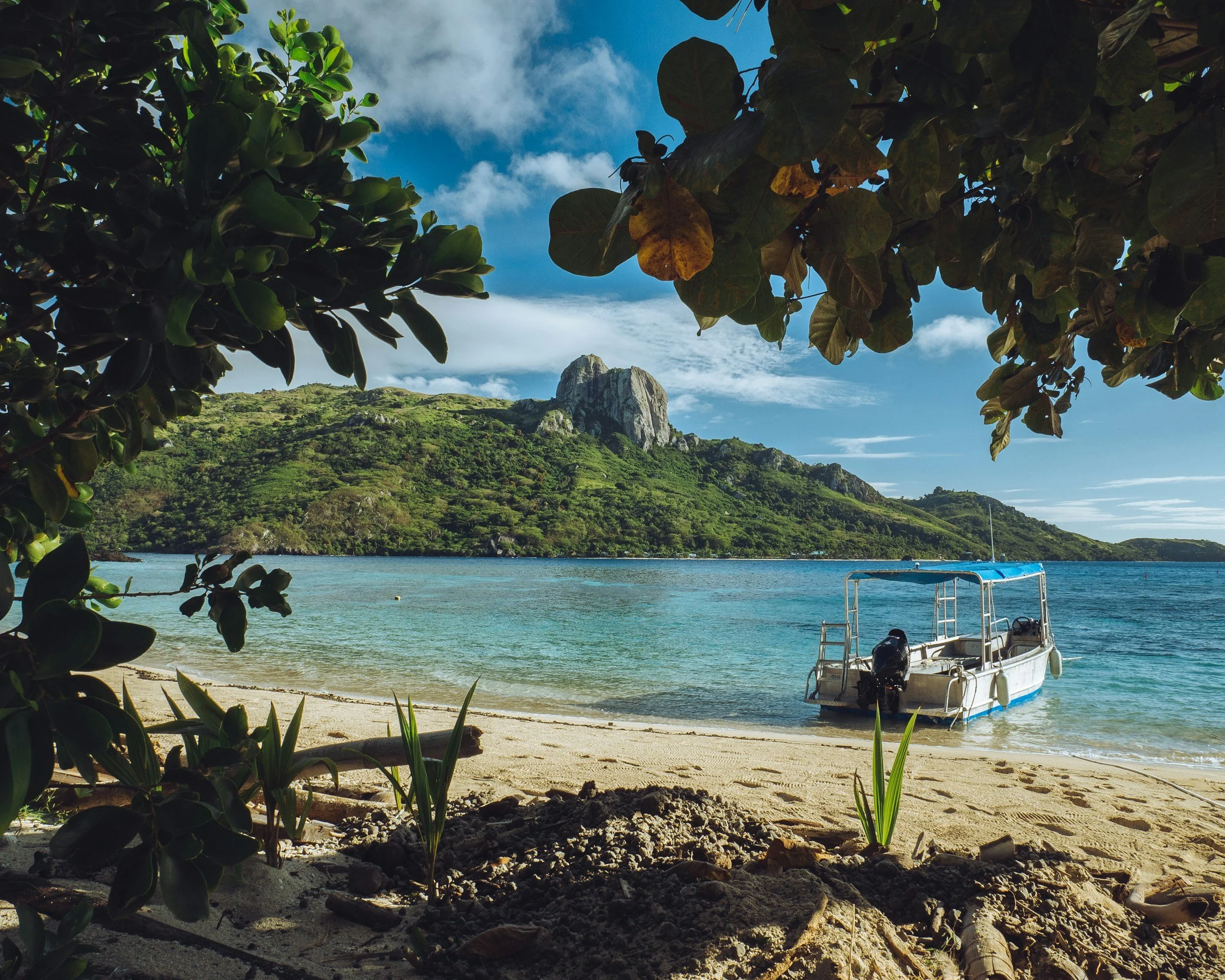 Tropical beach scene with a small boat anchored on the sandy shore, surrounded by lush green foliage, overlooking clear blue water and a distant, tree-covered mountain with a prominent peak under a partly cloudy sky.