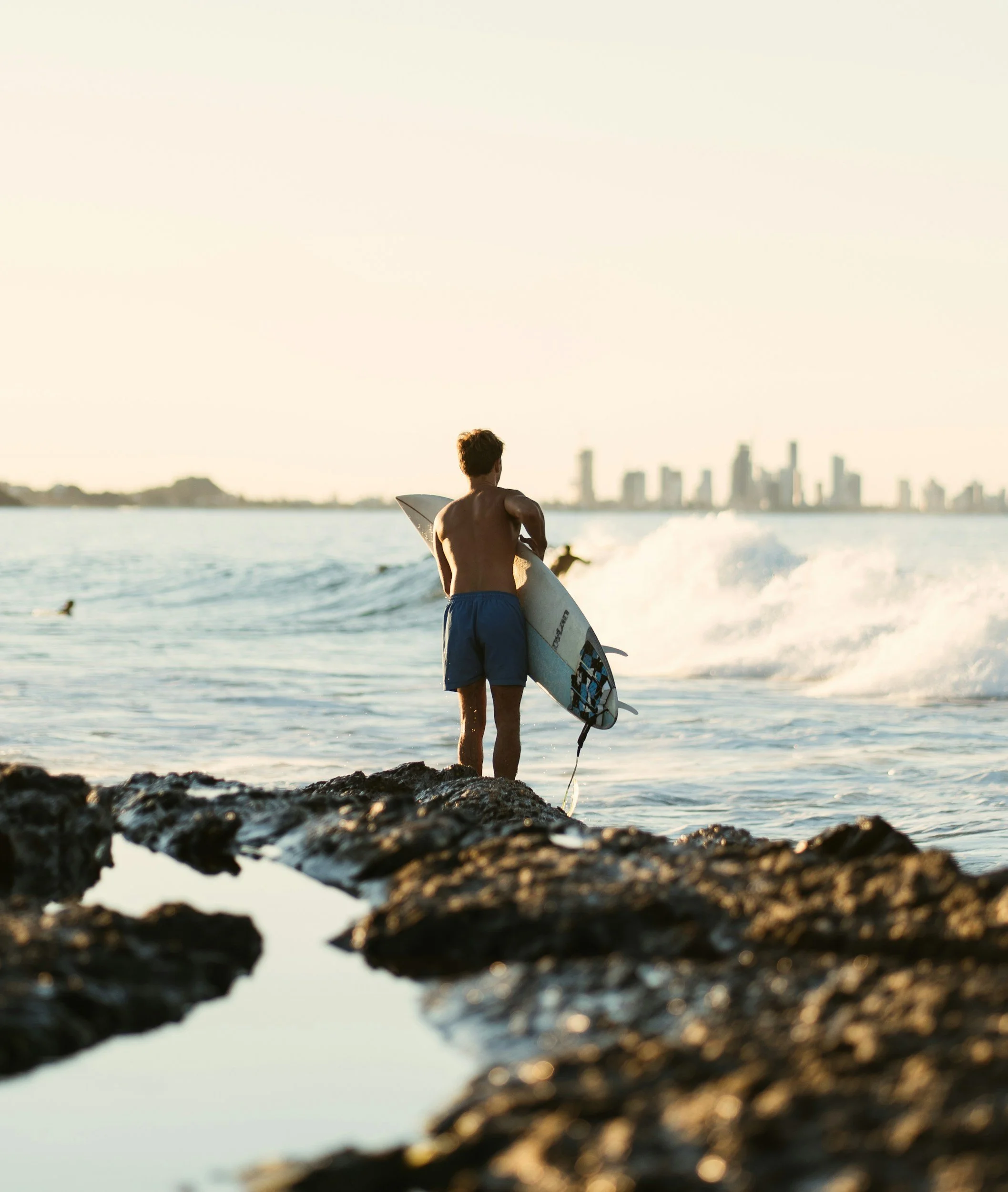 Surfer standing on rocks holding a surfboard, looking at the ocean waves, with city skyline in the background.