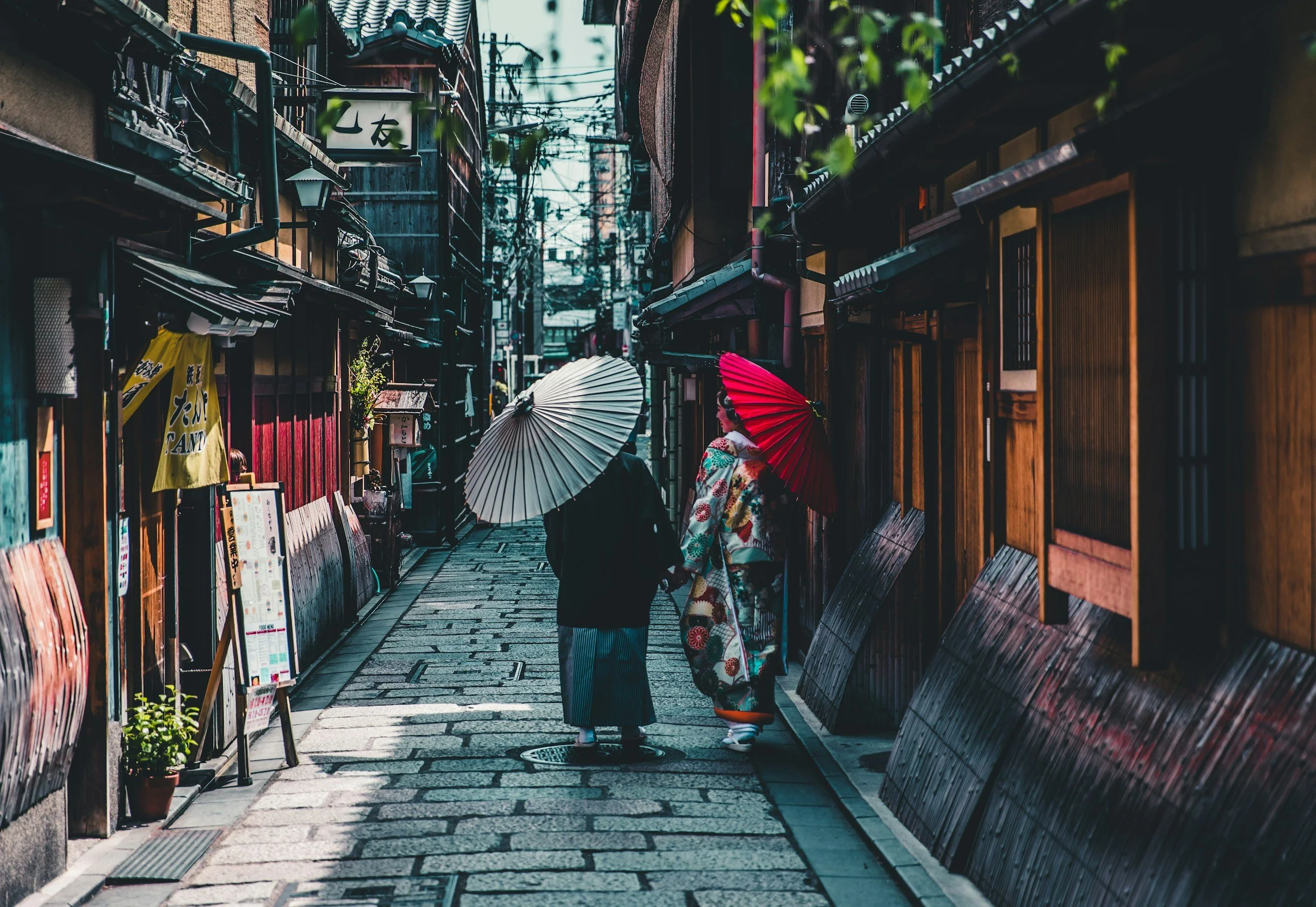 Traditional Japanese street with two people holding umbrellas, wearing kimonos, stone pathway, wooden buildings.
