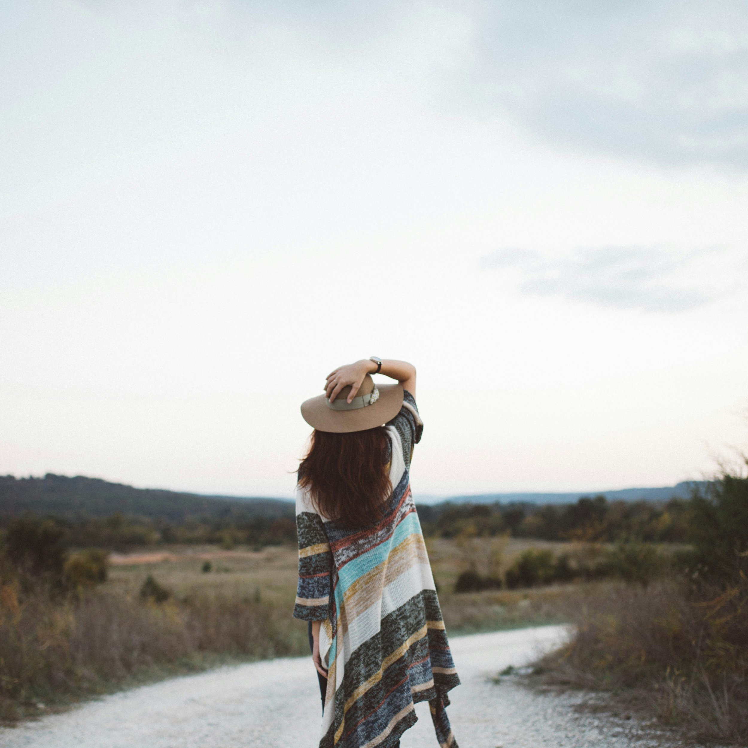 Woman standing on a countryside path, wearing a colorful poncho and a hat, looking at the landscape.