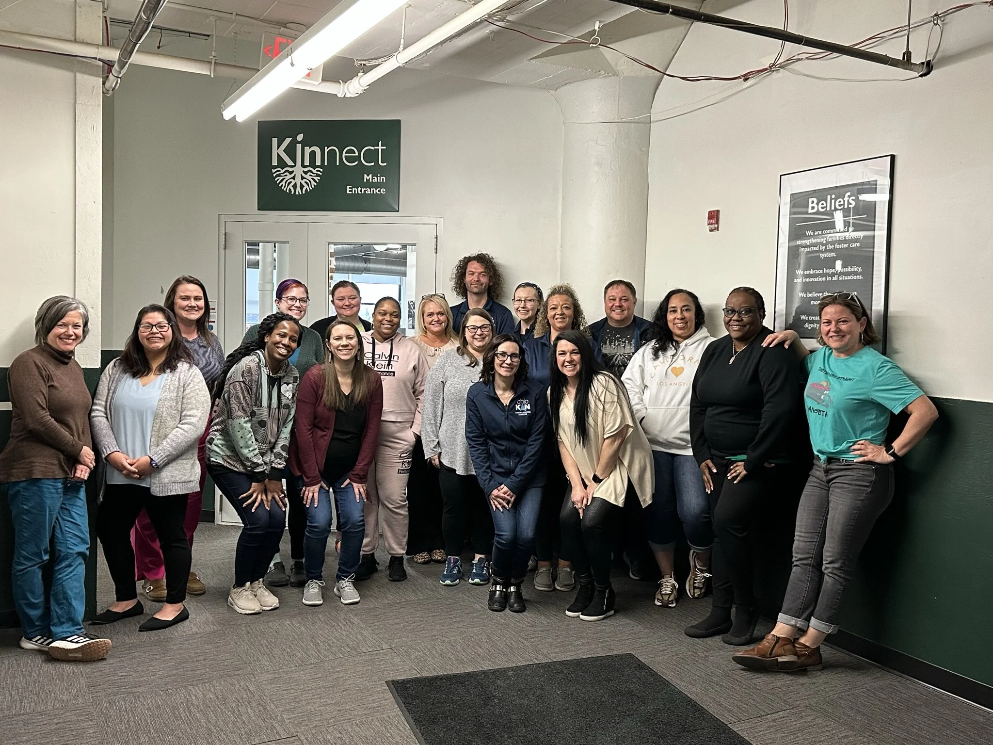 A group of diverse people standing and smiling in a hallway under a sign that says 'Kinnect Main Entrance.' There is a framed poster on the wall with text titled 'Beliefs.' They are casually dressed, suggesting a casual or friendly environment.