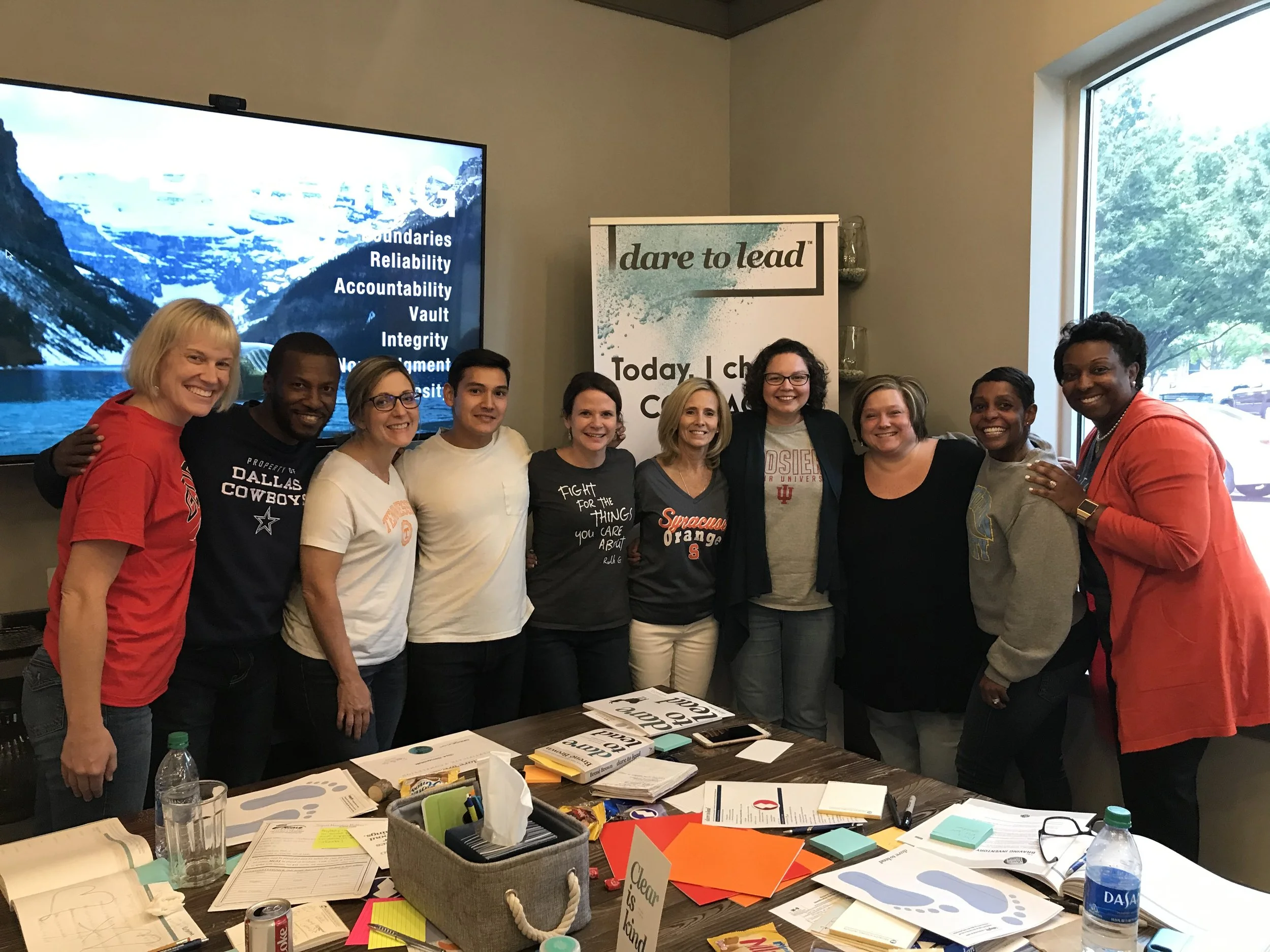 A group of nine people smiling and standing together indoors, with a banner in the background that reads "dare to lead." A table in front of them is cluttered with papers, books, and writing materials. The setting appears to be a collaborative or wor
