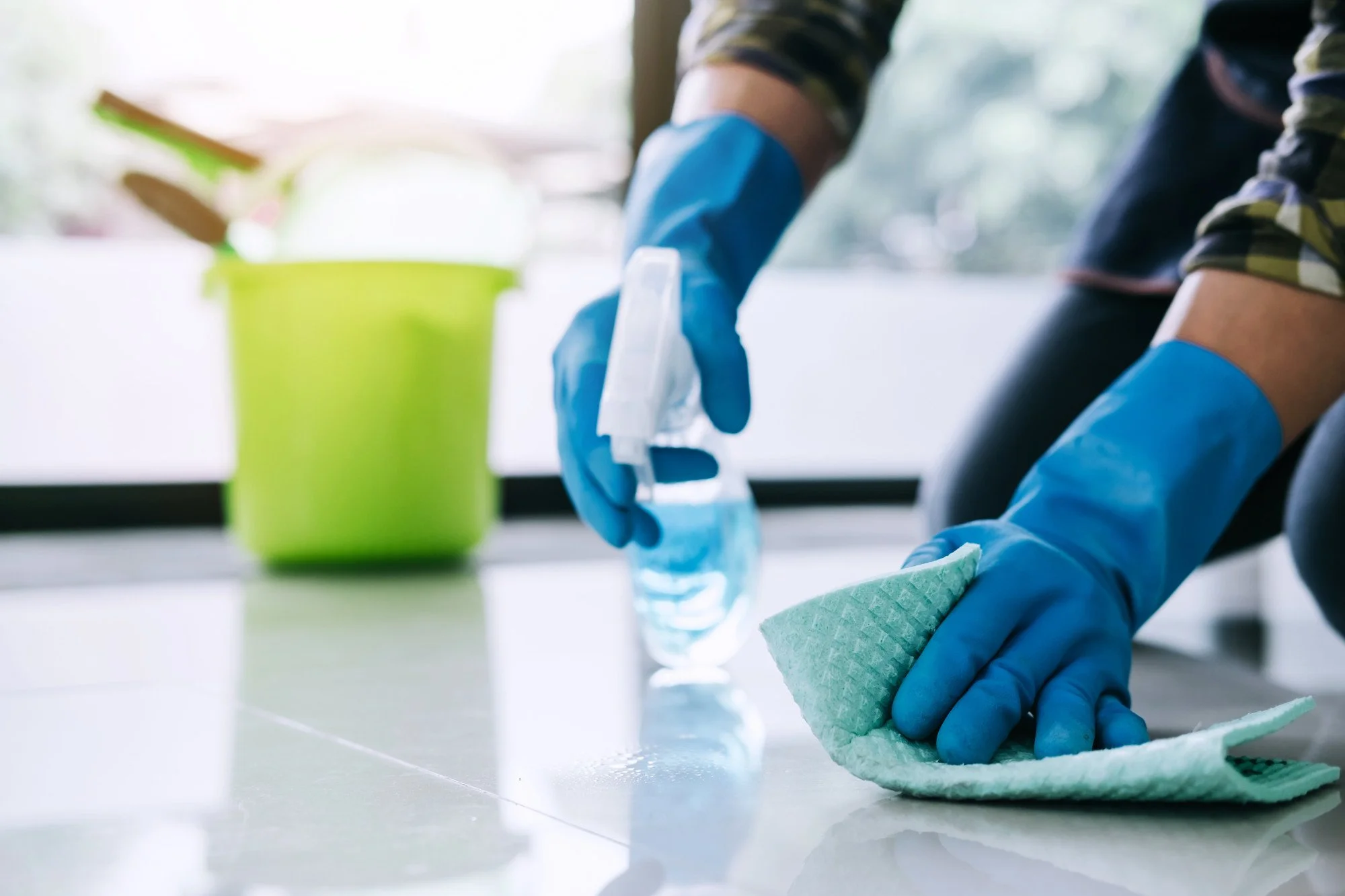 Person cleaning floor with blue gloves, spray bottle, and cloth; green bucket in background.