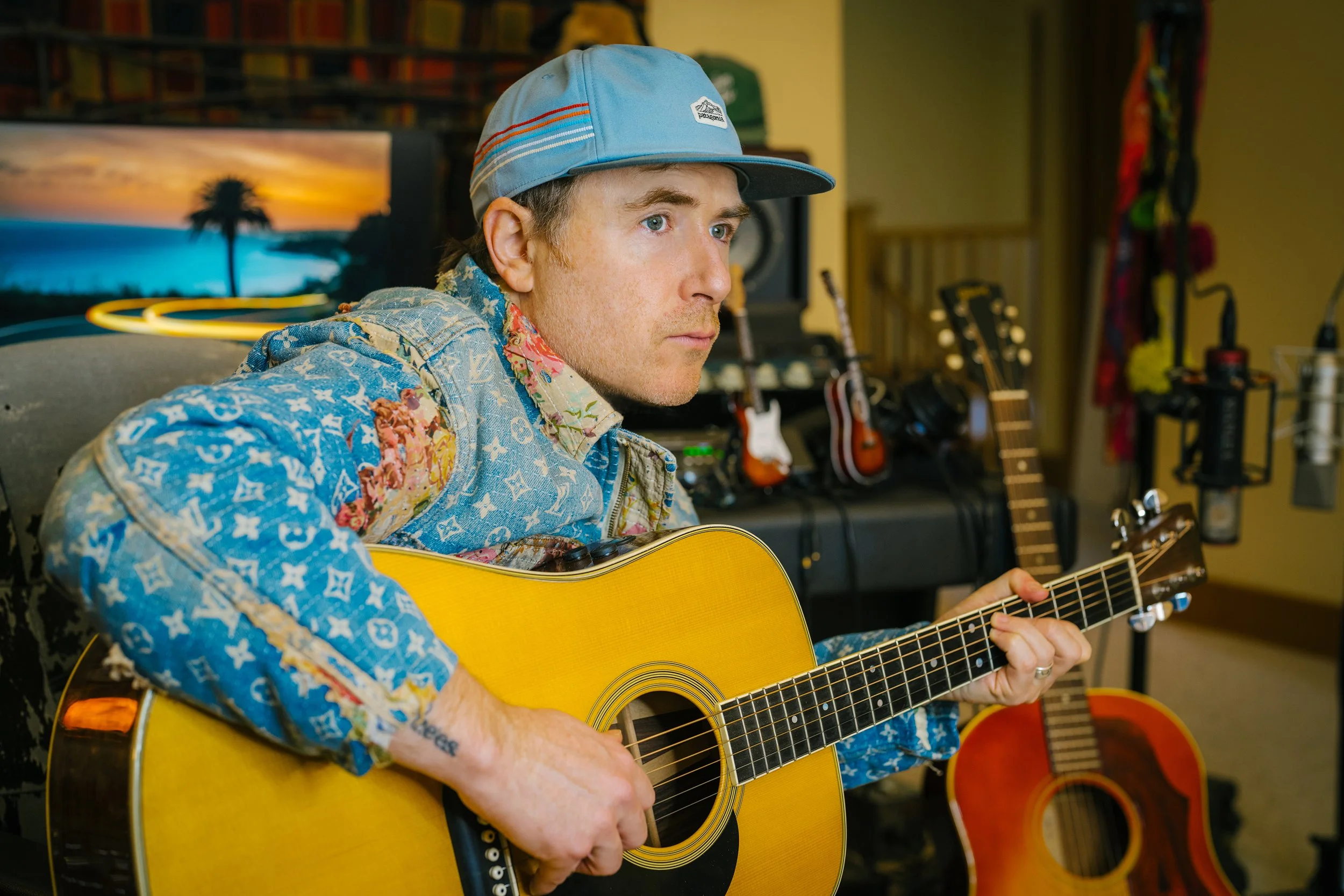 A man wearing a colorful, patterned denim jacket and a blue cap playing an acoustic guitar in a music studio filled with other guitars and recording equipment.