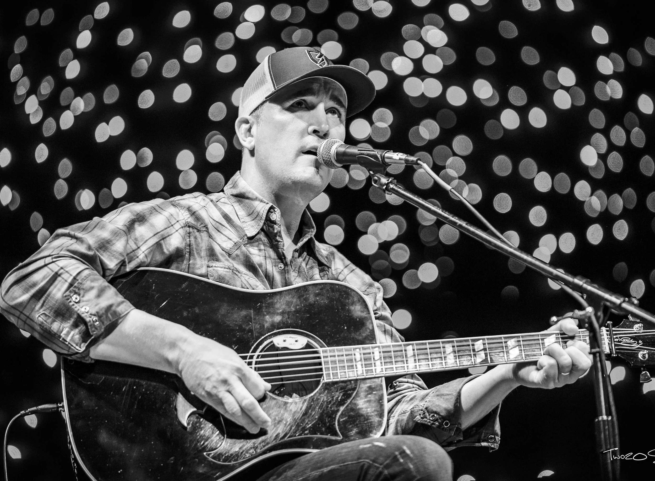 A man wearing a baseball cap and a plaid shirt is playing an acoustic guitar and singing into a microphone against a blurred background of circular lights.