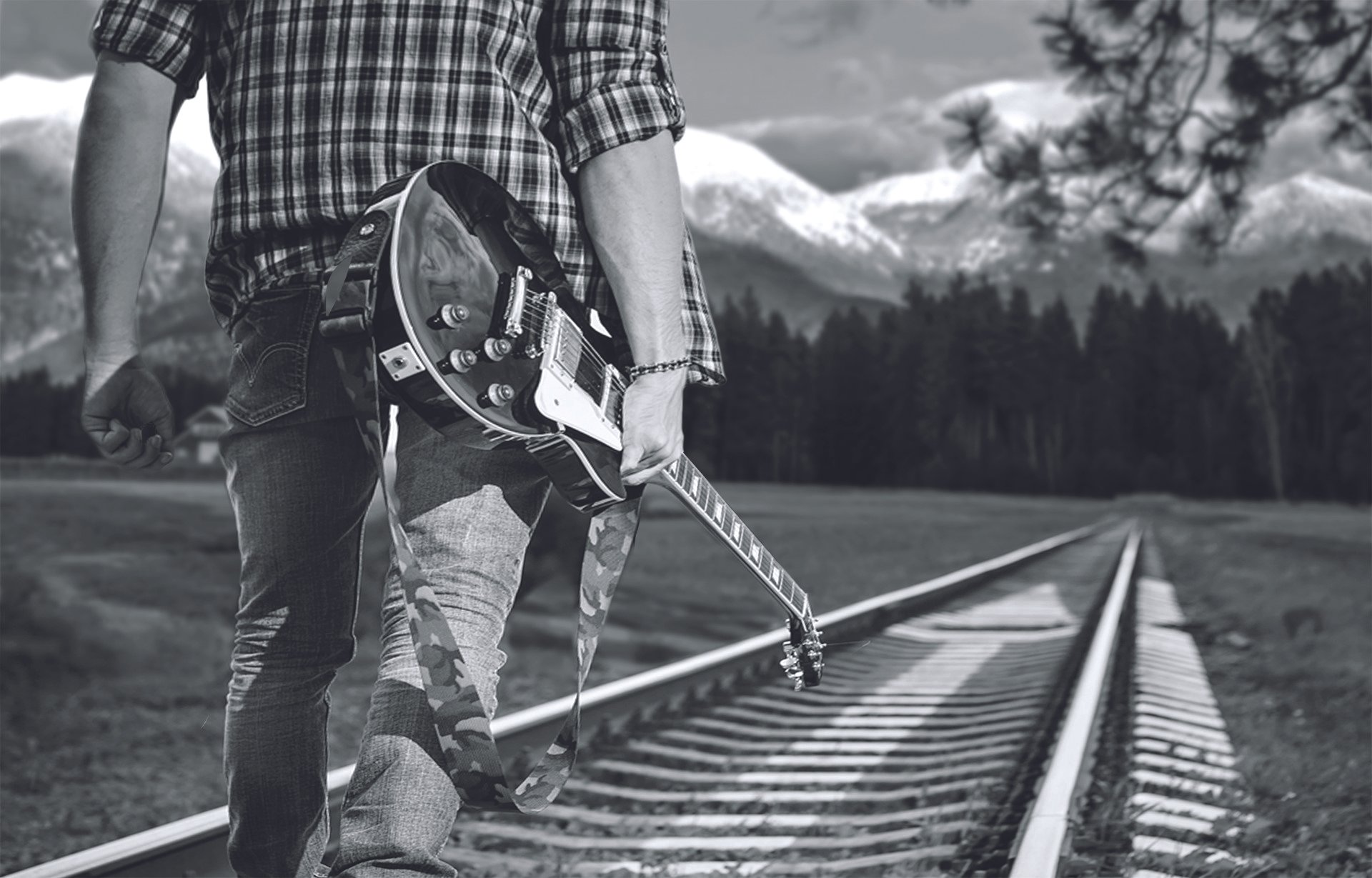 A person holding a guitar by their side standing on railway tracks with mountains and trees in the background, in black and white.