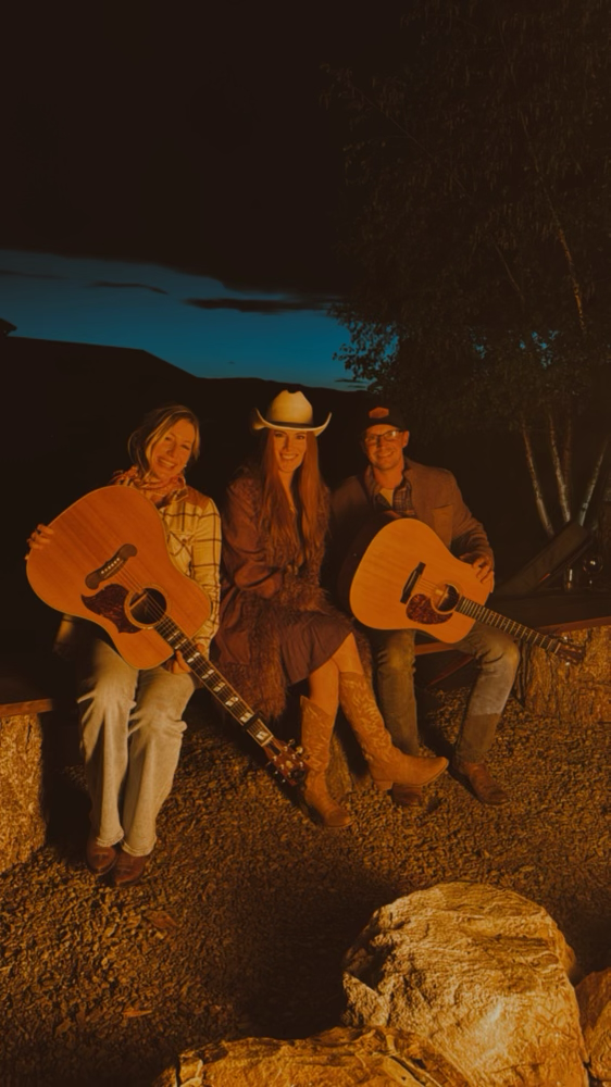 Three people sitting outdoors at night, illuminated by a campfire, with two holding acoustic guitars and wearing country-themed clothing and hats.