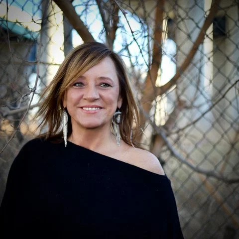 A woman with shoulder-length hair smiling, wearing a black off-shoulder top and earrings, standing outdoors near a fence and leafless trees.