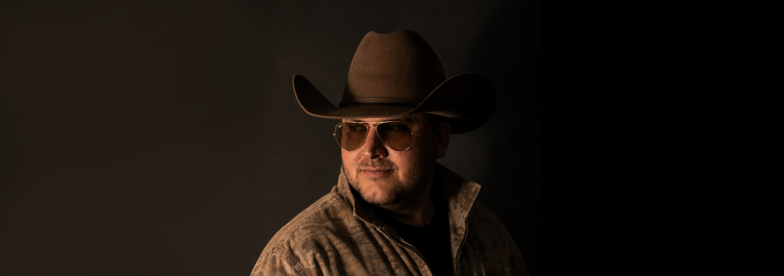 A man wearing a large cowboy hat, sunglasses, and a camouflage jacket, looking to the side against a dark background.