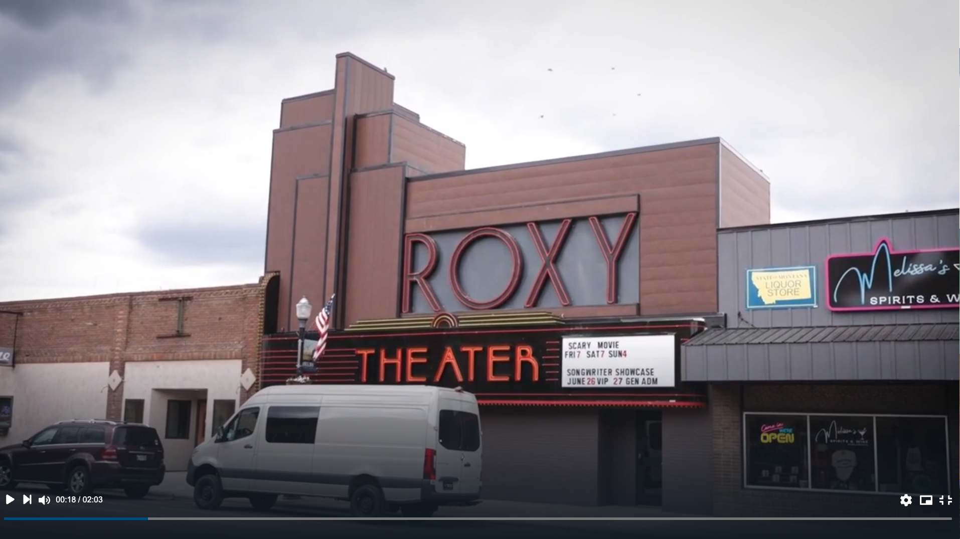 The Roxy Theater building with a large vertical sign displaying 'ROXY' and a smaller sign below reading 'THEATER.' Adjacent to the theater, there are signs for a liquor store and spirits and wine. There are cars parked in front, one is a white van. The sky above is overcast.